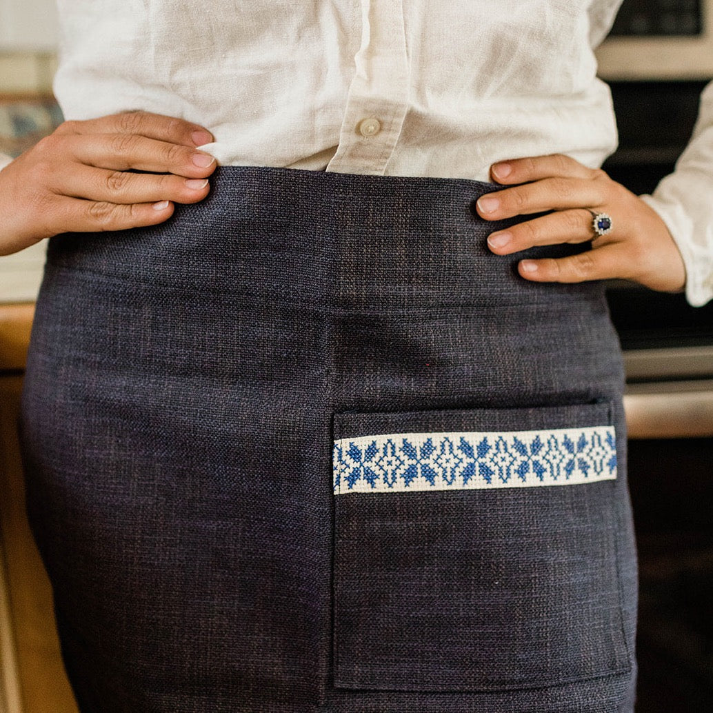 A person stands in a kitchen, hands on hips, wearing the Farah Half Apron - Blue over a white shirt and dark skirt with a blue patterned pocket.