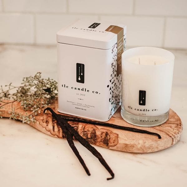 A Hibernate - Patchouli Soy Candle in a white glass jar, accompanied by an elegant decorative tin, rests on a wooden board alongside vanilla beans and dried flowers. The ensemble is presented on a marble surface, with the gentle fragrance of patchouli imparting an earthy touch to this tranquil setting.