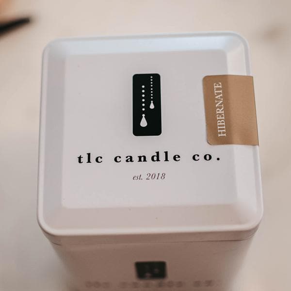 A Hibernate - Patchouli Soy Candle in a white glass jar, accompanied by an elegant decorative tin, rests on a wooden board alongside vanilla beans and dried flowers. The ensemble is presented on a marble surface, with the gentle fragrance of patchouli imparting an earthy touch to this tranquil setting.