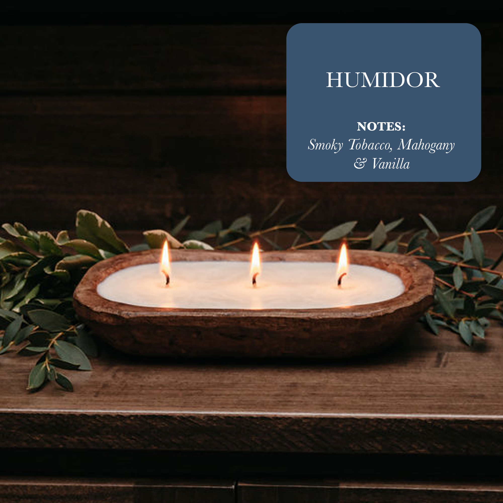 A 3-Wick Dough Bowl Soy Candle named "Humidor" flickers on a table, enveloped by lush greenery. The text highlights scent notes of smoky tobacco, mahogany, and vanilla. This wooden bowl candle creates a cozy ambiance with its rich and inviting aroma.