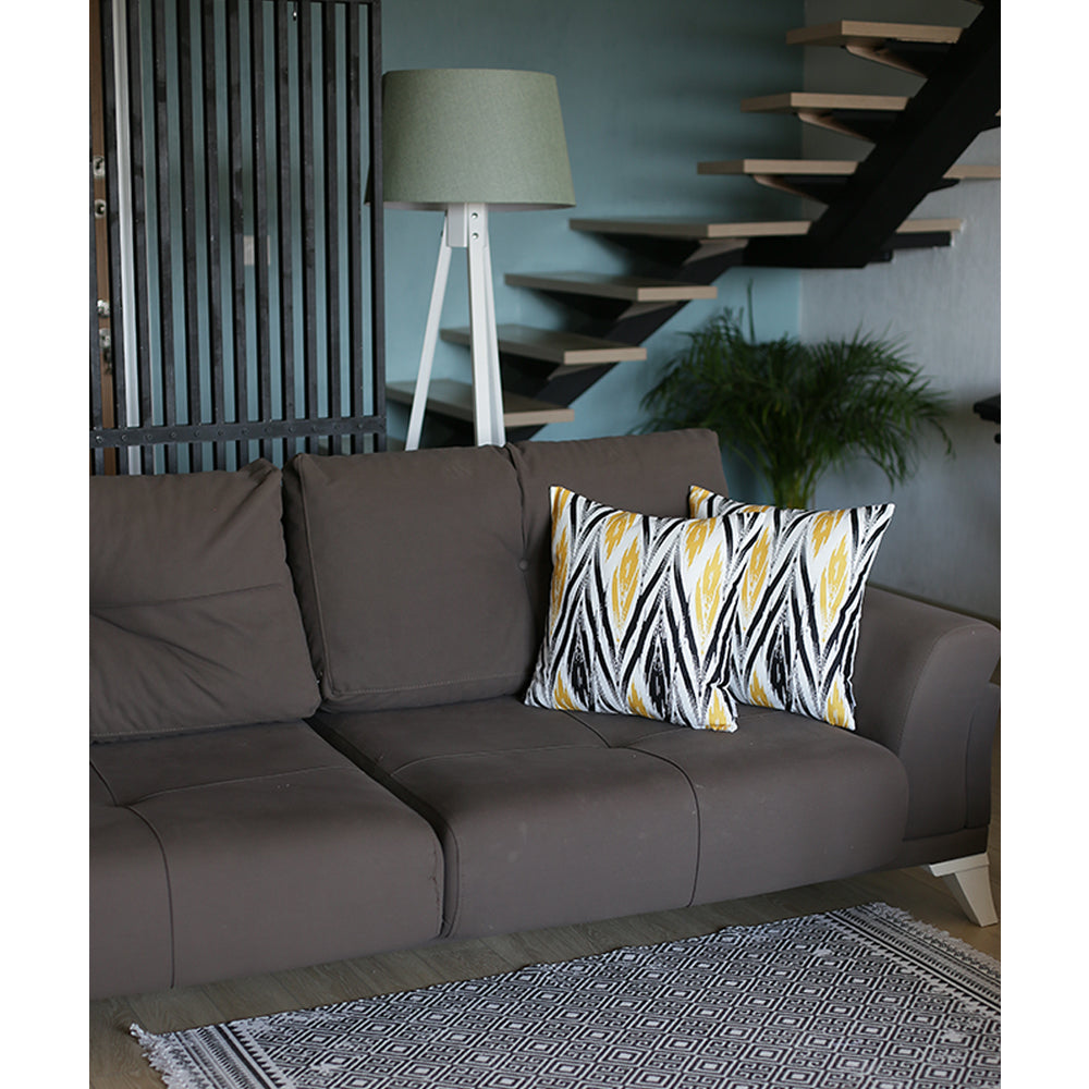 A gray sofa with Geometric Yellow Lit Square 18" Throw Pillow Covers sits in front of a tall lamp. A potted plant is near a stairway with floating steps, and a patterned rug graces the floor.