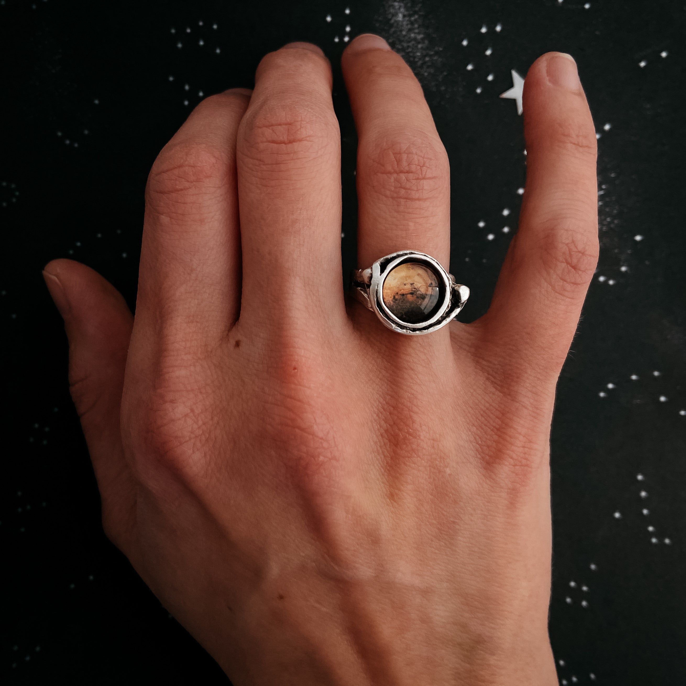 A hand wears the Mars and Moons Ring, featuring a silver tone with a spherical, galaxy-like design reminiscent of space exploration jewelry, set against a starry background.