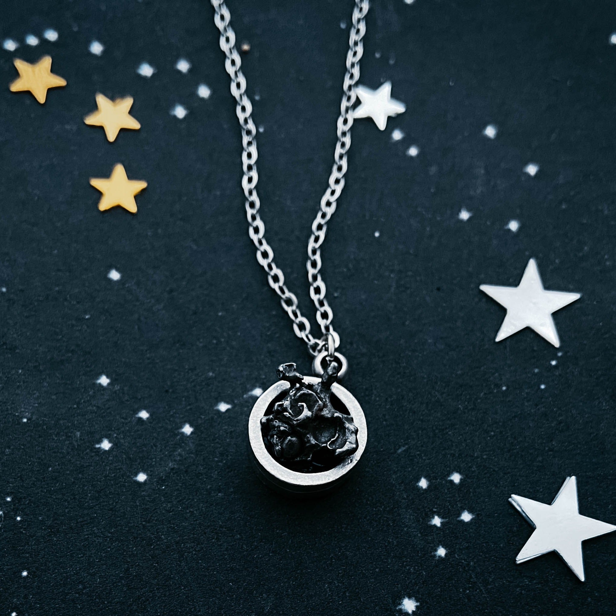 A Small Round Meteorite Pendant Necklace in Matte Brushed Silver rests on a dark background, surrounded by scattered gold and silver star-shaped decorations, embodying the enchanting allure of outer space jewelry.