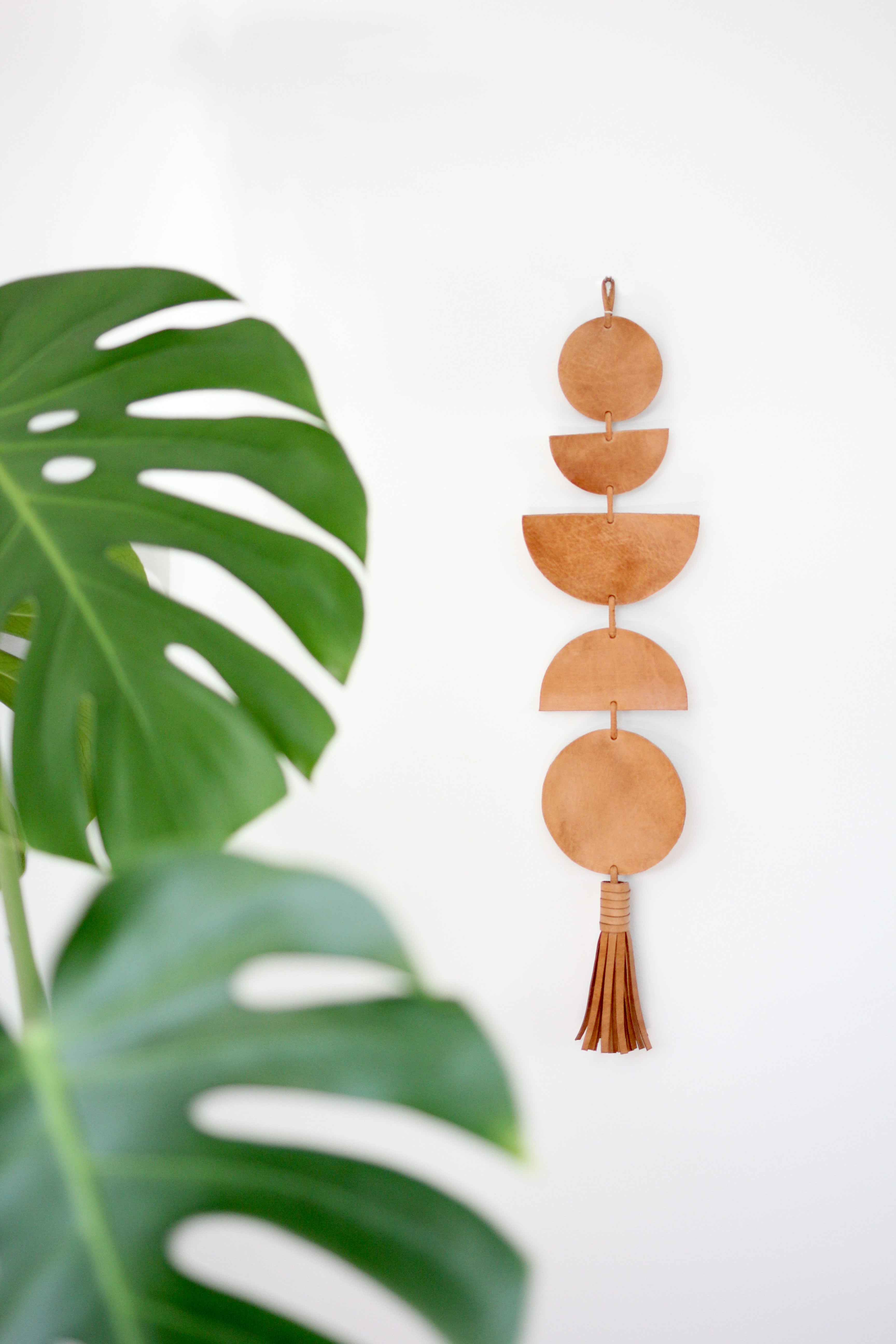 The Luna Moon Phase Wall Hanging, crafted with handmade circular and semicircular shapes from brown leather, is placed beside a large green monstera leaf on a white background, evoking the phases of the moon.