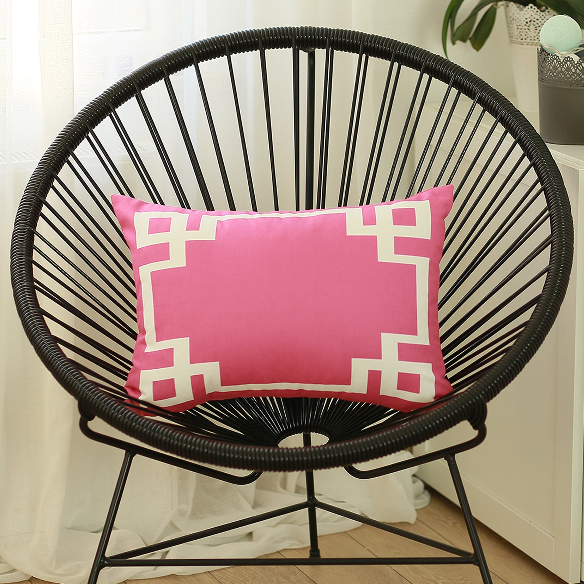 A round black wire chair, adorned with a Geometric Pink&White Square 18" Throw Pillow Cover & Insert, featuring high-quality digital printing, sits indoors on a wooden floor near a plant.