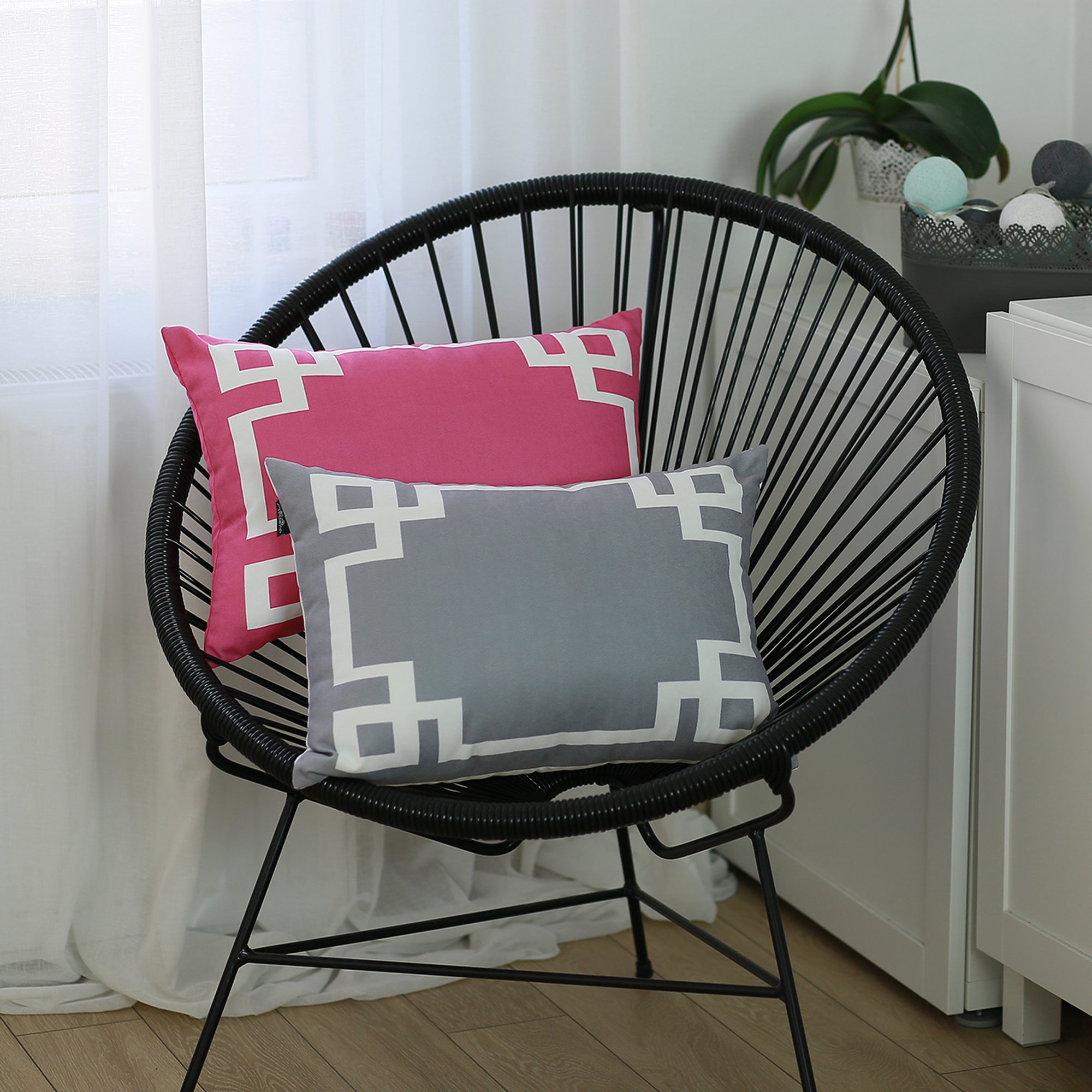 A round black wire chair, adorned with a Geometric Pink&White Square 18" Throw Pillow Cover & Insert, featuring high-quality digital printing, sits indoors on a wooden floor near a plant.