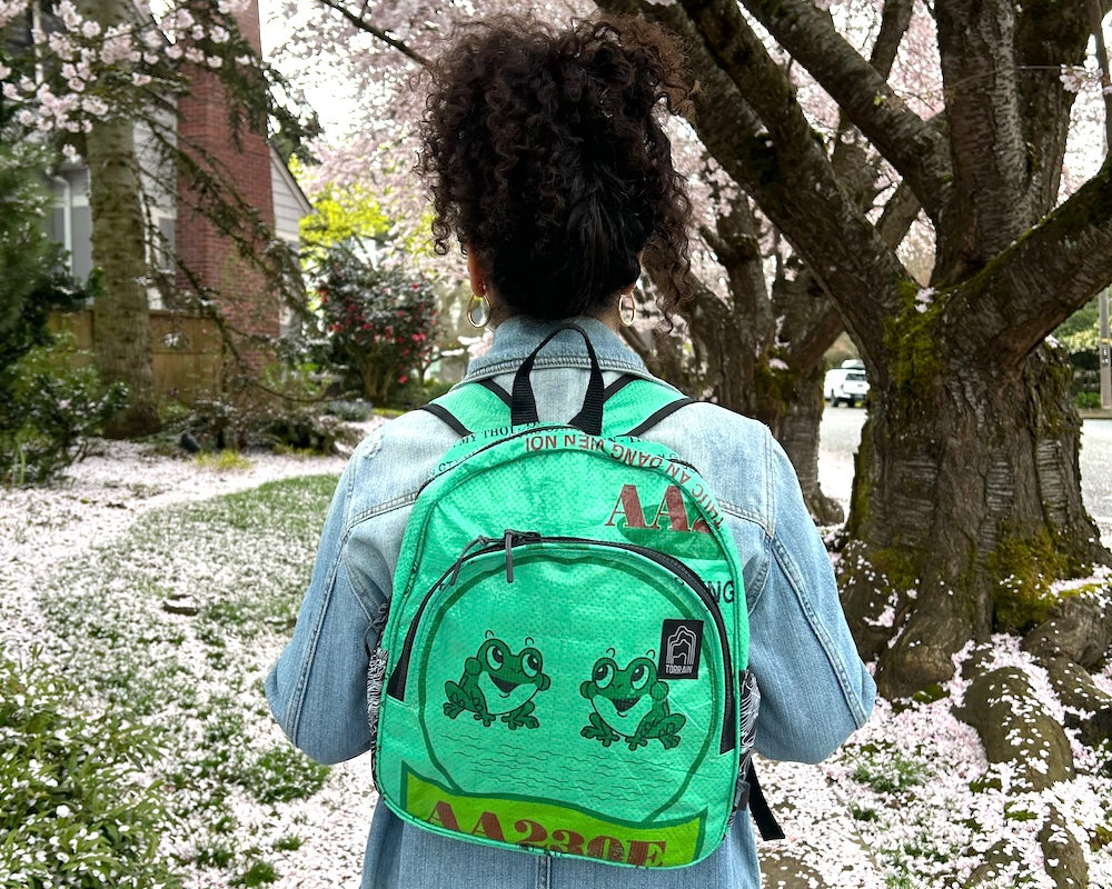 Introducing the Scout Backpack: a vividly designed accessory featuring a yellow front, an orange top, and blue patterned sides. Crafted from upcycled materials, it includes padded shoulder straps for comfort. The logo elegantly embellishes the front pocket, adding a stylish flair.