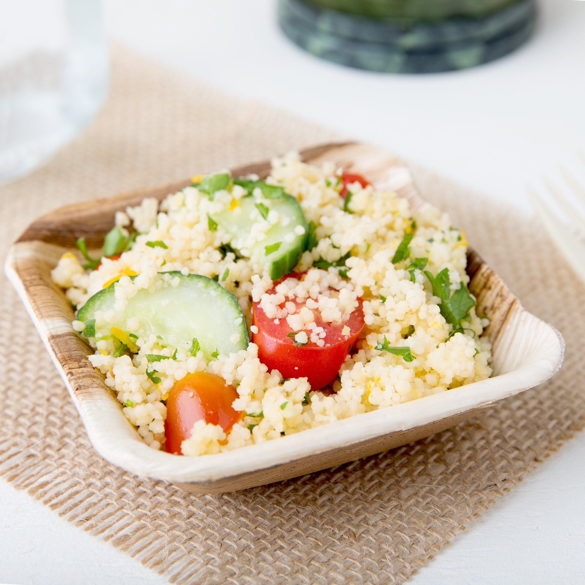 A bowl of couscous salad with cucumber, tomato, and parsley is elegantly presented on a burlap mat, served in eco-friendly Palm Leaf Square Bowl 3.5" Inch - 3oz Mini (25 count).