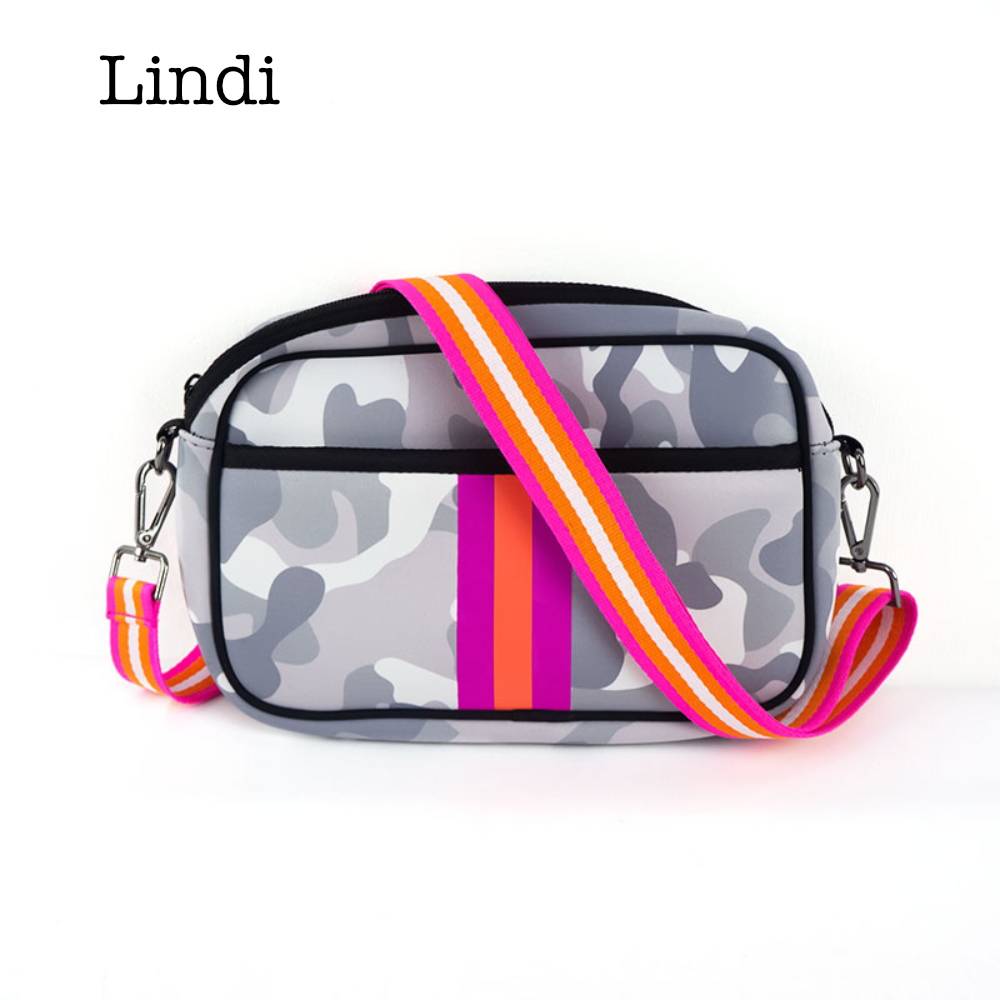 A person in a white dress carries the Neoprene Compact Crossbody, which features a camouflage-patterned fabric highlighted with pink and orange stripes.