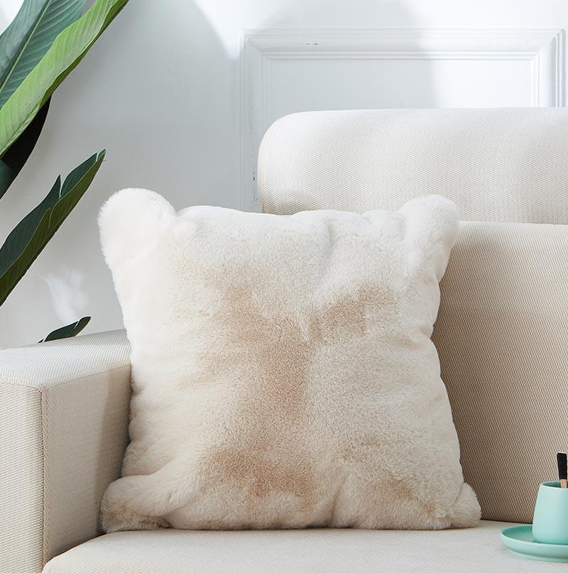A fluffy white Agnes Luxury Beige Chinchilla Faux Fur Pillow on a cream sofa, with a green plant and white wall in the background.