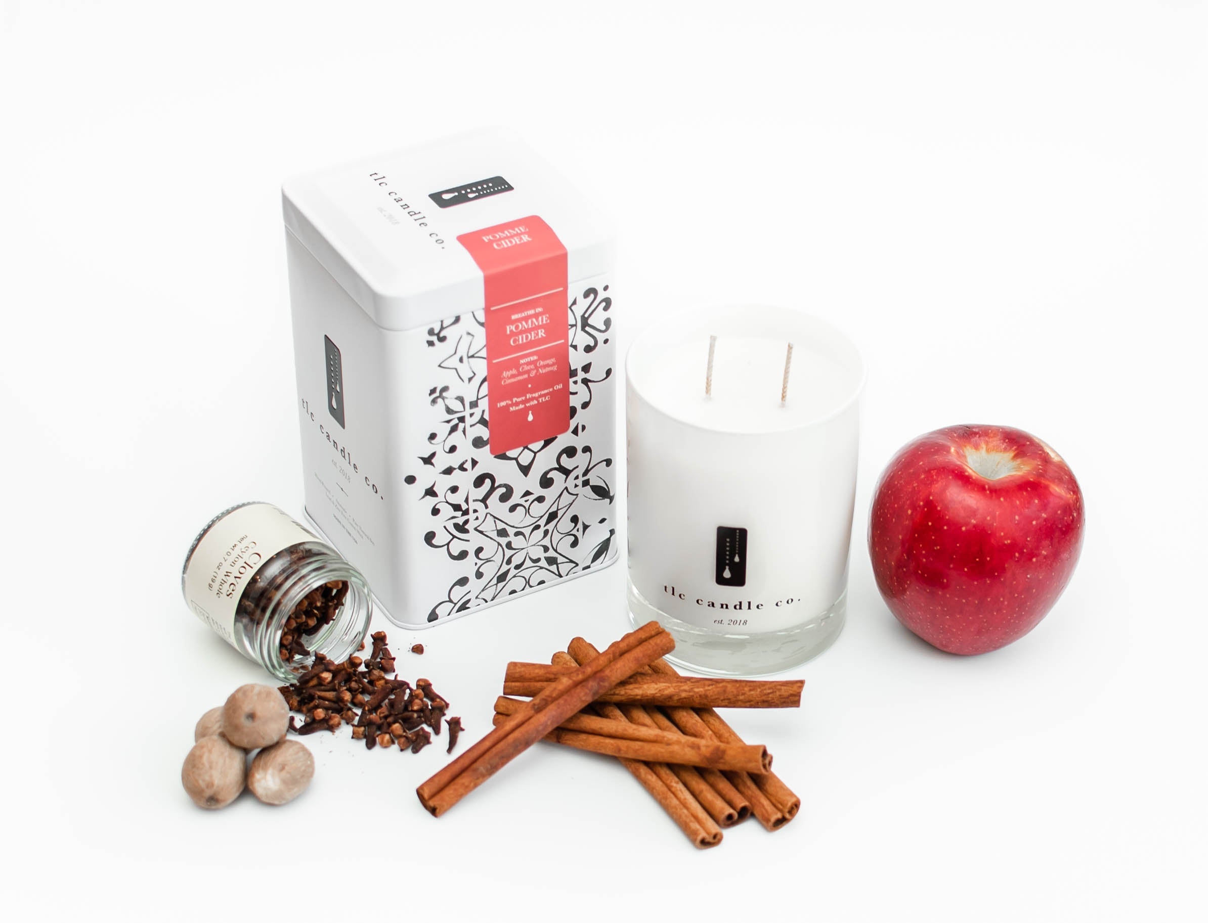 The Pomme Cider soy candle jar, accompanied by a decorative box, cinnamon sticks, nutmeg, cloves, and a red apple, is artfully arranged on a white surface.