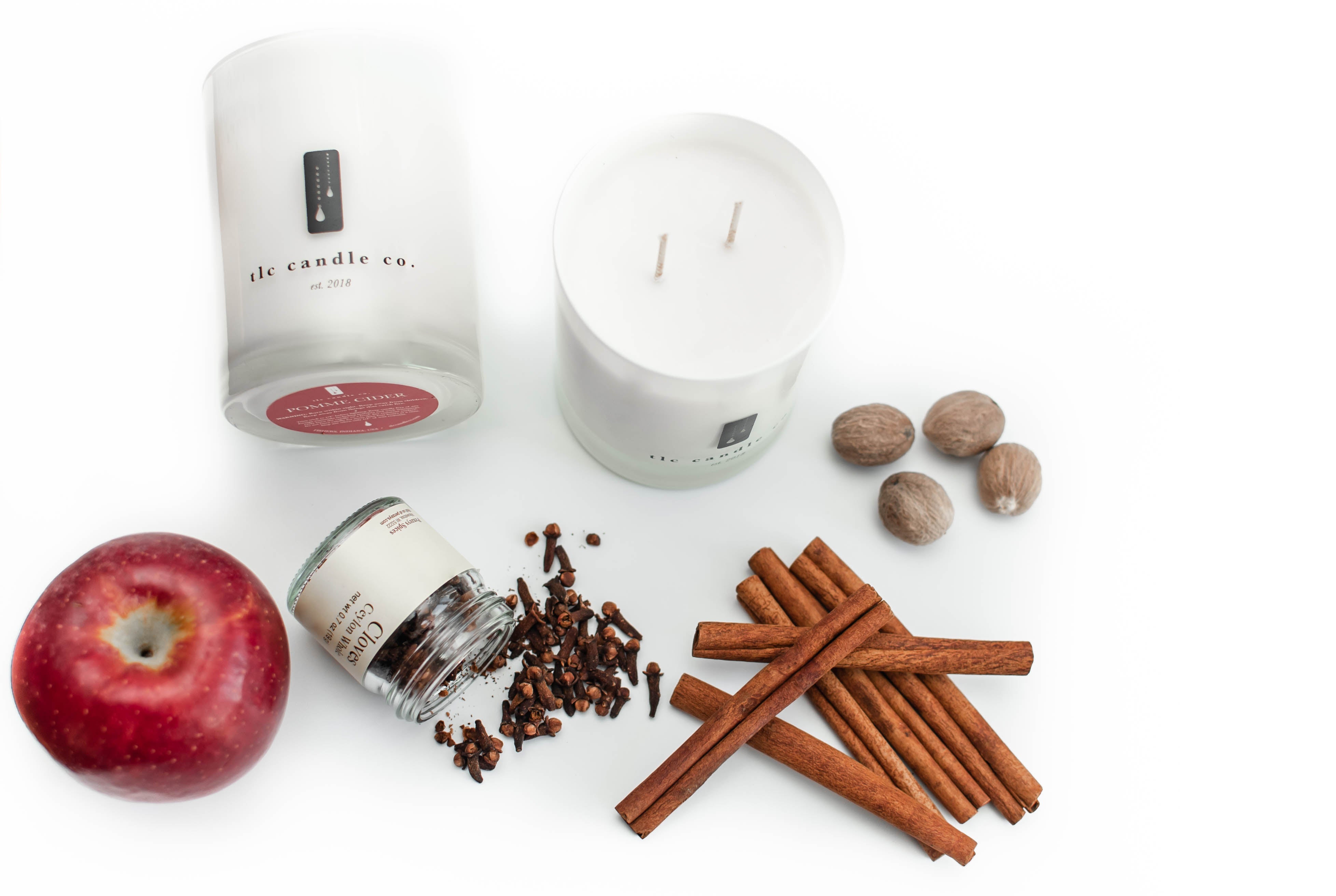 The Pomme Cider soy candle jar, accompanied by a decorative box, cinnamon sticks, nutmeg, cloves, and a red apple, is artfully arranged on a white surface.