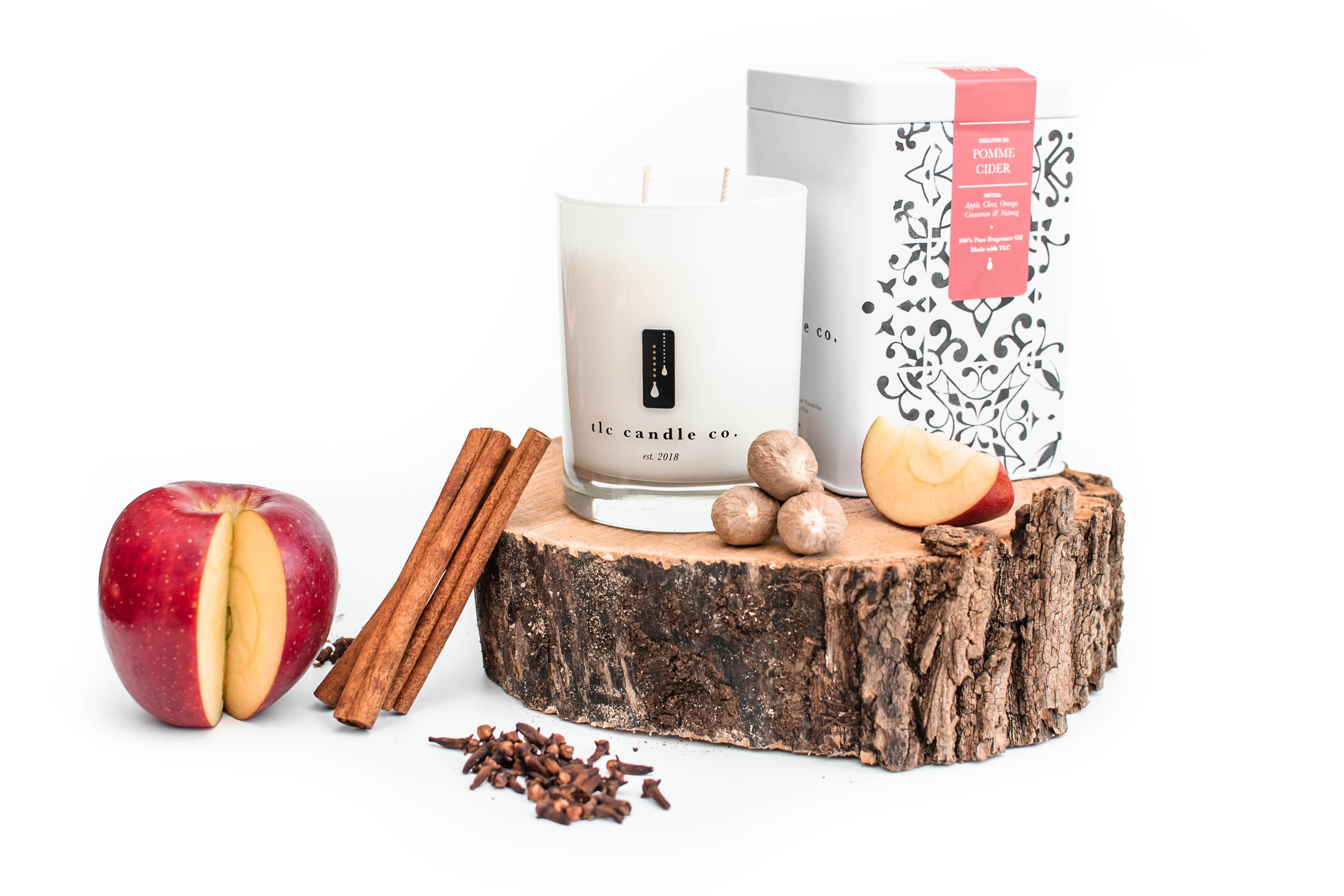 The Pomme Cider soy candle jar, accompanied by a decorative box, cinnamon sticks, nutmeg, cloves, and a red apple, is artfully arranged on a white surface.