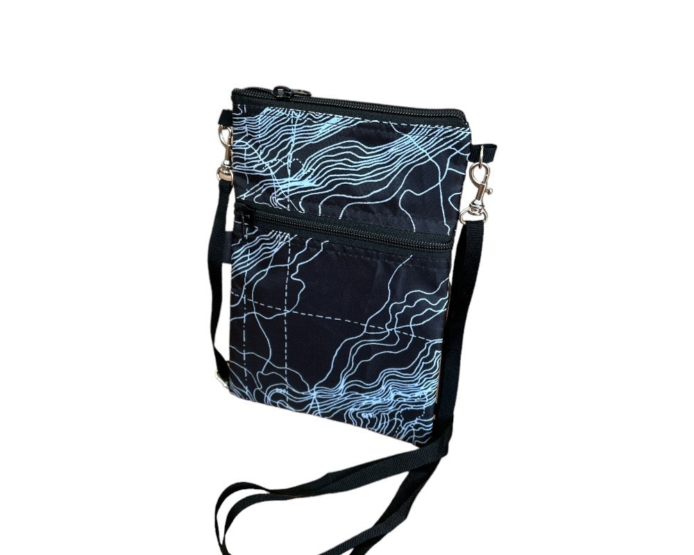 The Breezy Crossbody showcases a vibrant blue fish print on a pink background, crafted from upcycled materials. It includes two black zippered compartments and an adjustable black strap.