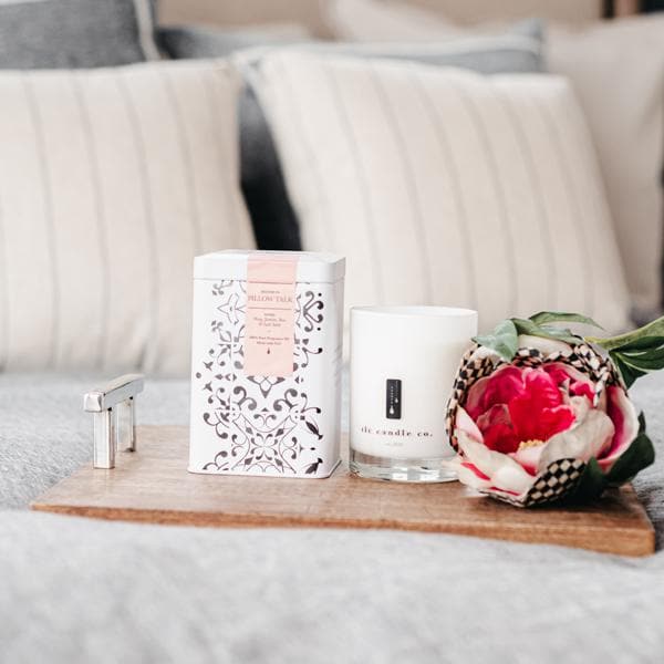 A decorative tin, a lighter, and the Pillow Talk - Peony Soy Candle are arranged on a wooden tray, resting on a soft grey surface with striped pillows in the background.