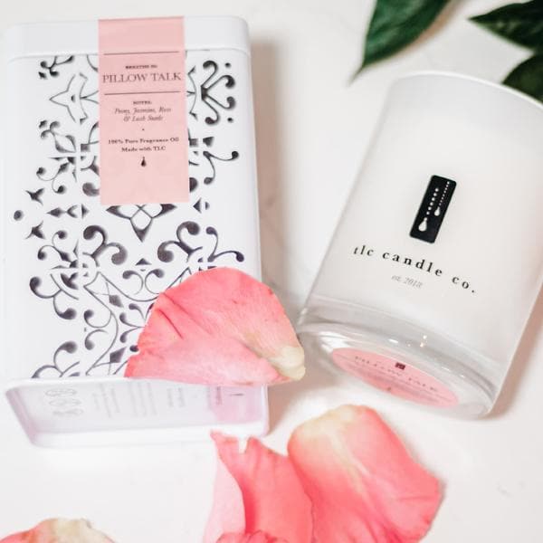 A decorative tin, a lighter, and the Pillow Talk - Peony Soy Candle are arranged on a wooden tray, resting on a soft grey surface with striped pillows in the background.