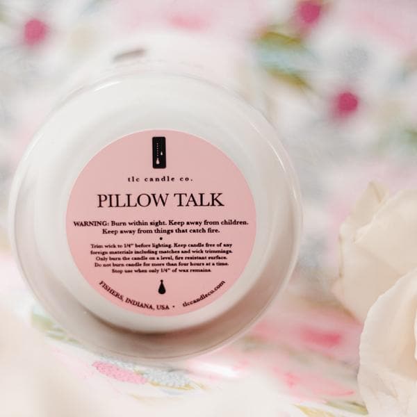 A decorative tin, a lighter, and the Pillow Talk - Peony Soy Candle are arranged on a wooden tray, resting on a soft grey surface with striped pillows in the background.