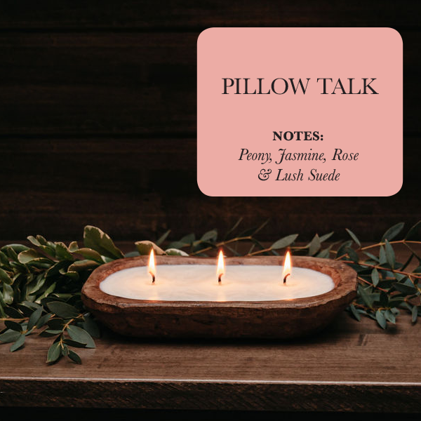 A three-wick dough bowl soy candle named "Pillow Talk" rests on a wooden surface, surrounded by lush green foliage and delicate rose petals. The scent features notes of peony, jasmine, rose, and lush suede.