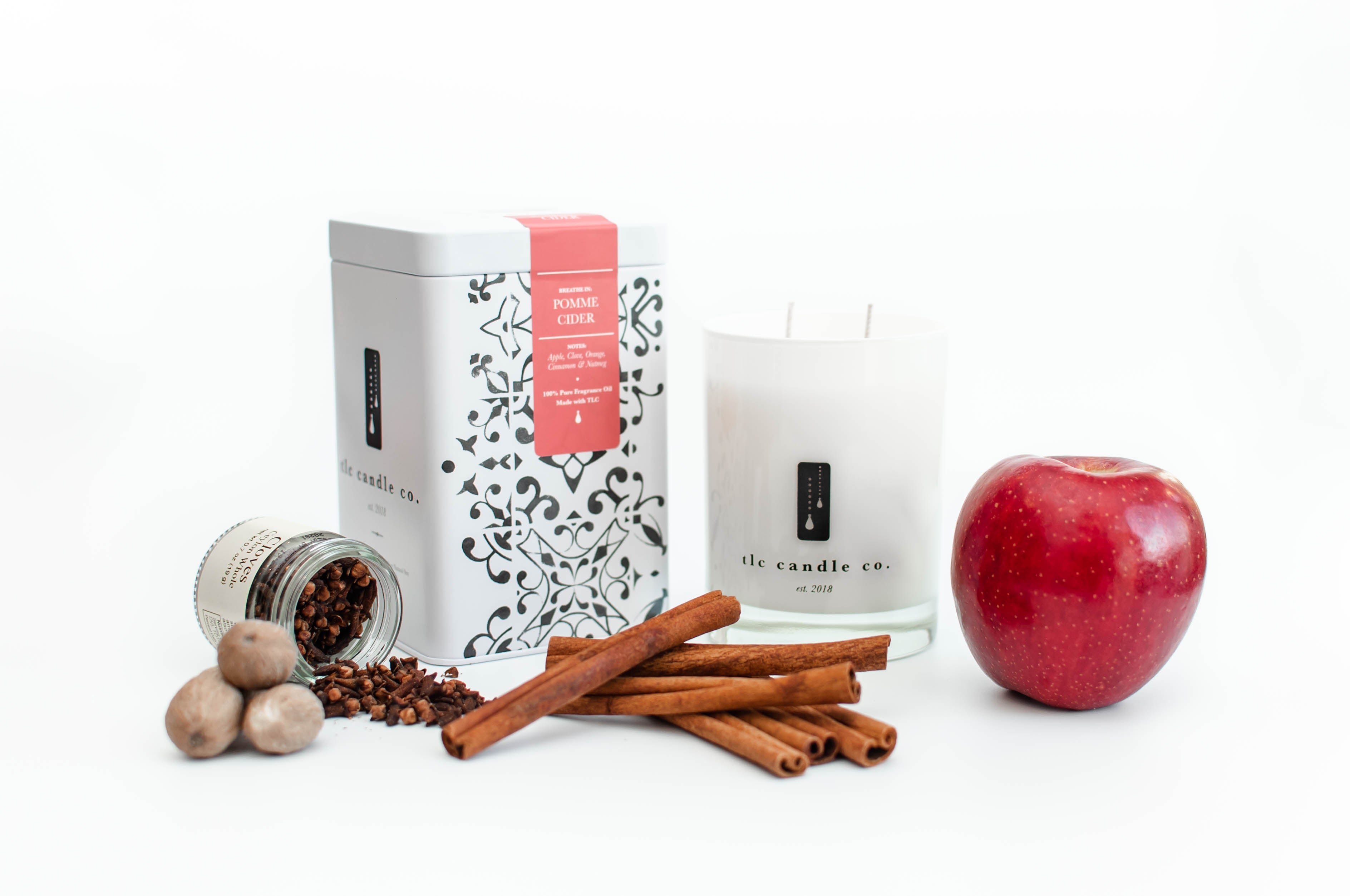 The Pomme Cider soy candle jar, accompanied by a decorative box, cinnamon sticks, nutmeg, cloves, and a red apple, is artfully arranged on a white surface.