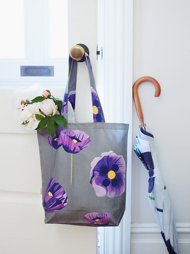 A stylish "Tote Bag: Purple Poppies on Grey," crafted from cotton canvas, hangs on a doorknob, showcasing its beautiful purple poppy design while holding white flowers. Nearby, an umbrella with a wooden handle leans against the door.