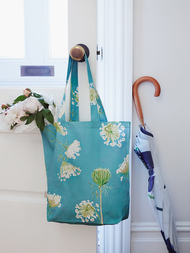 A cotton canvas tote bag featuring a Queen Anne's Lace pattern on a teal background hangs stylishly on a doorknob, next to a folded umbrella. White peonies rest gently on the door, adding an elegant touch to the scene.