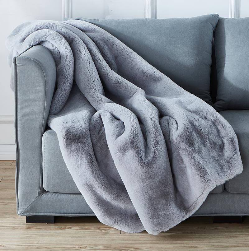 A stain-resistant Cassilda Luxury Grey Chinchilla Faux Fur Throw Blanket draped over a gray sofa in a room with wooden flooring.