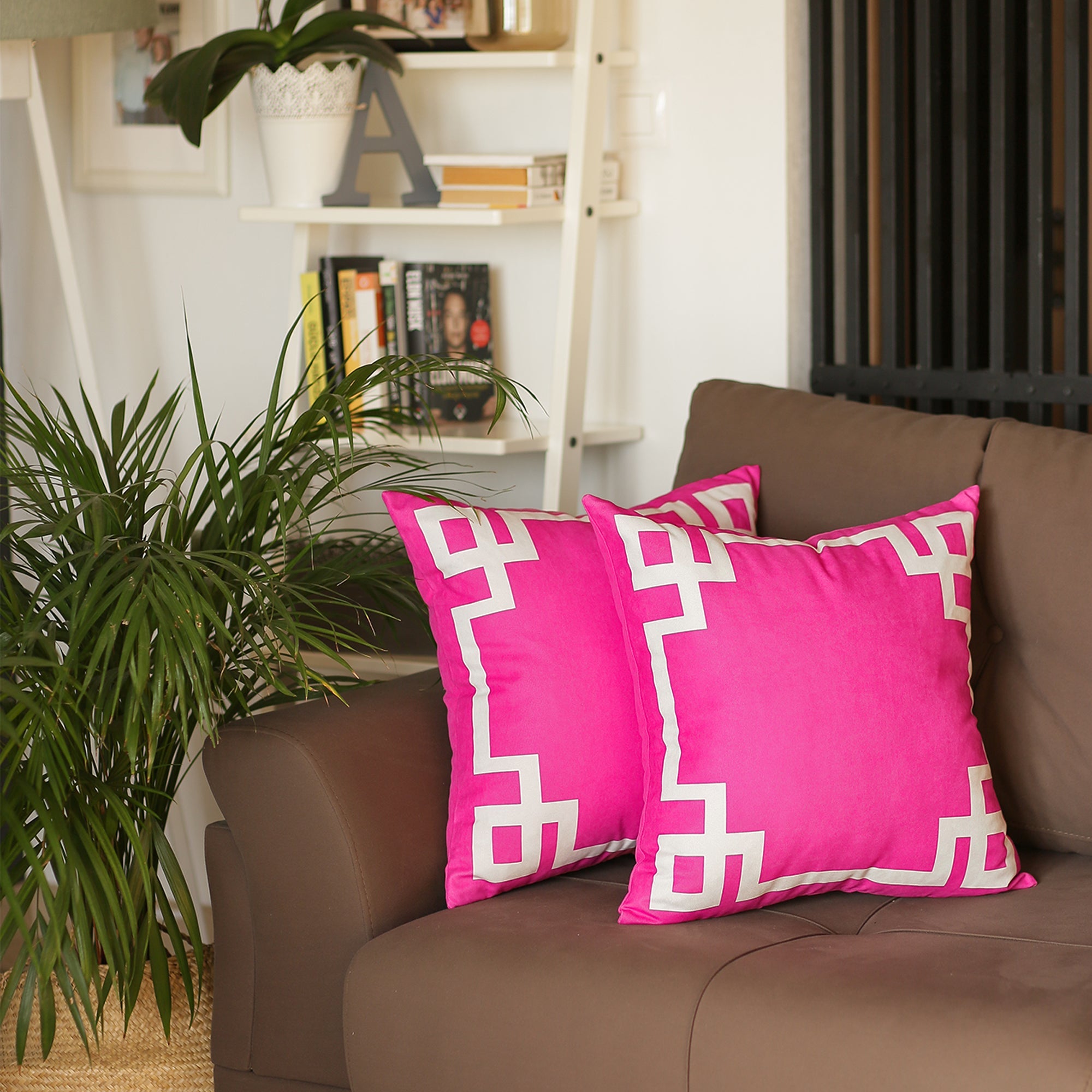 Adorning a brown sofa are two Geometric Pink&White Square 18" Throw Pillow Covers, each showcasing stylish geometric patterns. Paired with a green potted plant and a white bookshelf, these covers include convenient hidden zipper closures for effortless upkeep.