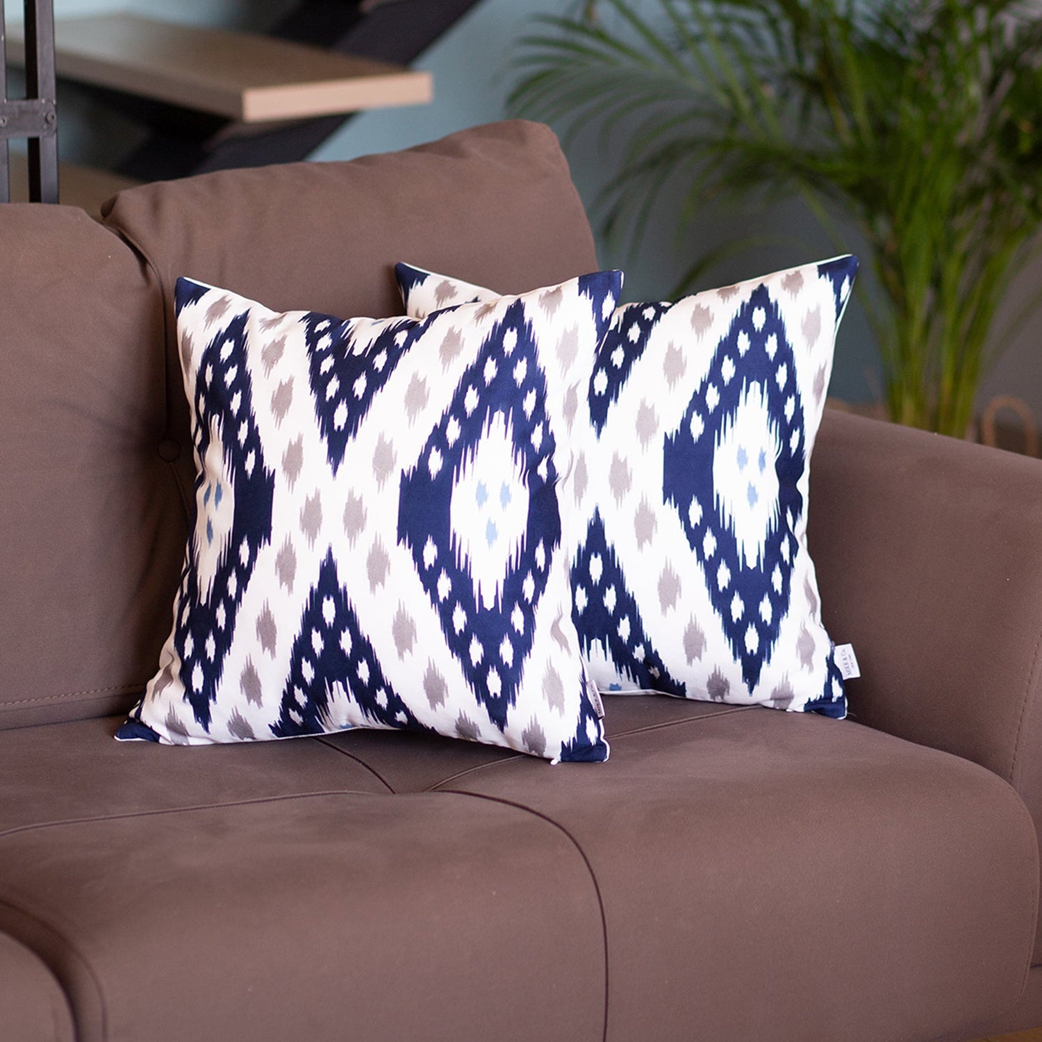 The brown sofa is adorned with the Ikat Set of 2 Square 18”x18” Boho Throw Pillow Covers, featuring a blue and white geometric pattern and hidden zipper closures for a seamless look. In the background, a plant peeks through, adding a touch of nature to the cozy space.