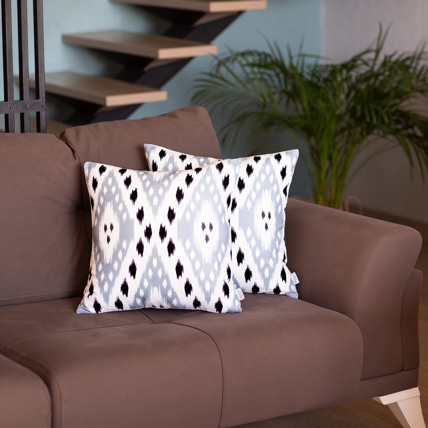 A pair of pillow covers from the Ikat Set of 2 Square 18’’x18’’ Boho Throw Pillow Covers, showcasing a blue and white ikat design with hidden zipper closures, are placed on a brown sofa. In the background, you can see a potted plant and a staircase.
