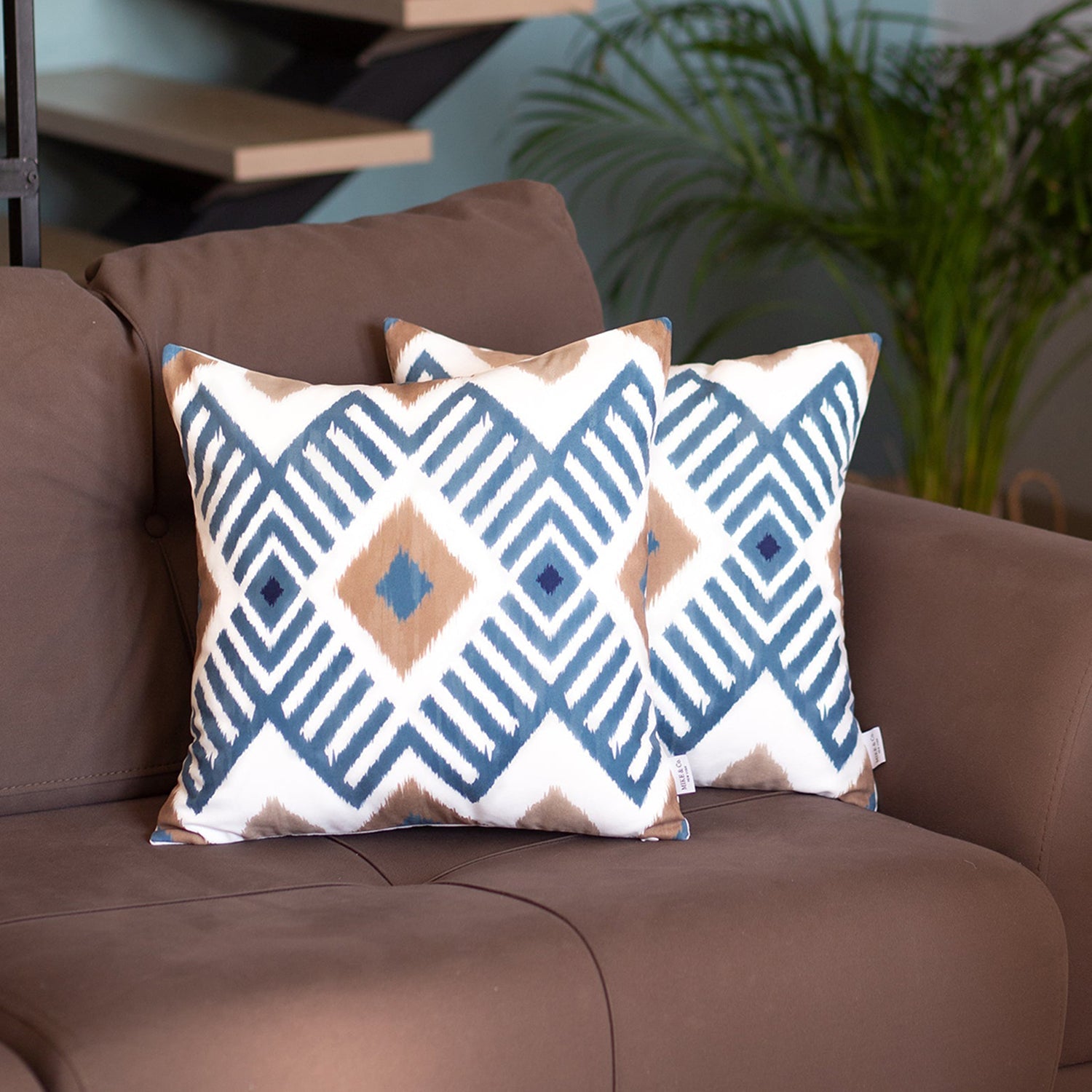 Two Ikat Set of 2 Square 18’’x18’’ Boho Throw Pillow Covers with geometric designs adorn a brown sofa, featuring hidden zipper closures. A vibrant green plant creates a serene backdrop, adding a touch of nature to the setting.