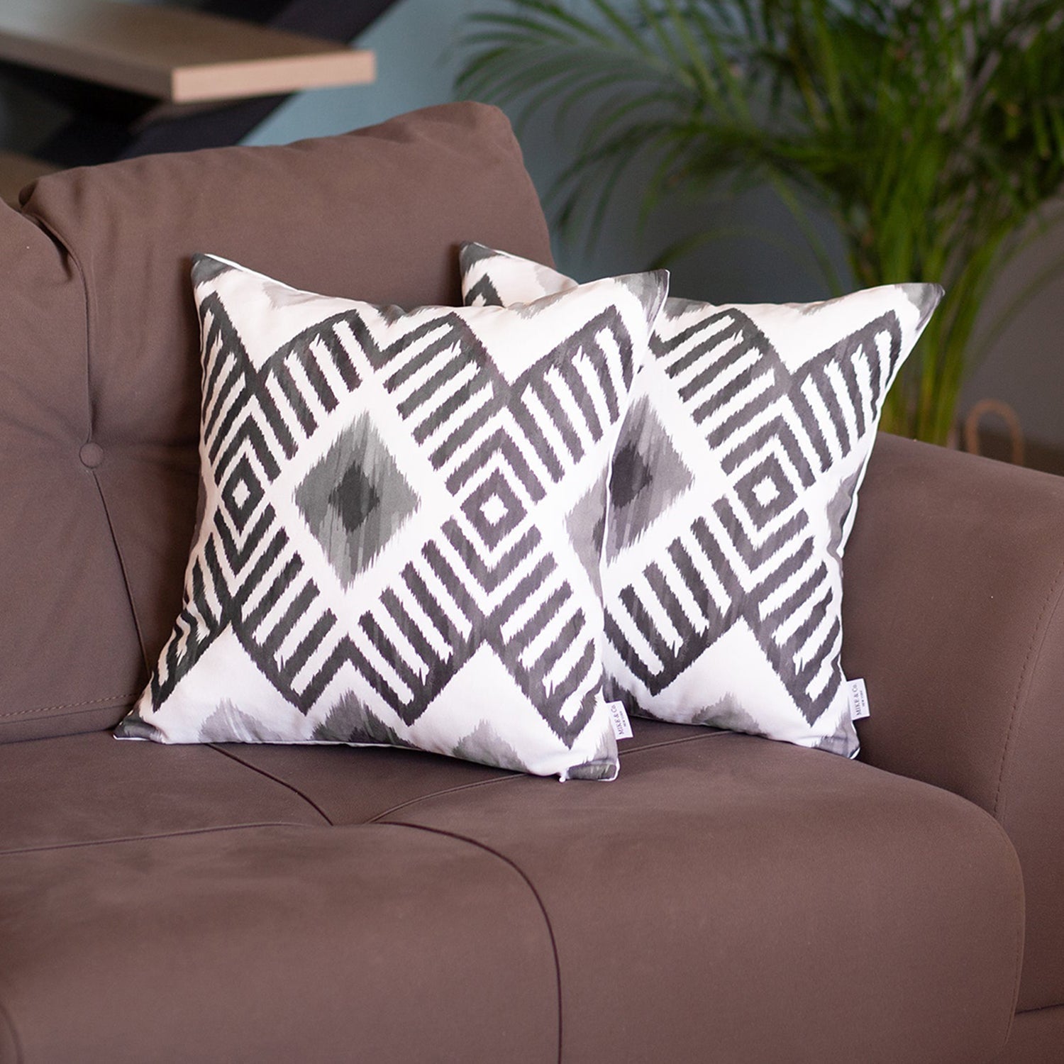 Two Ikat Set of 2 Square 18’’x18’’ Boho Throw Pillow Covers with geometric designs adorn a brown sofa, featuring hidden zipper closures. A vibrant green plant creates a serene backdrop, adding a touch of nature to the setting.