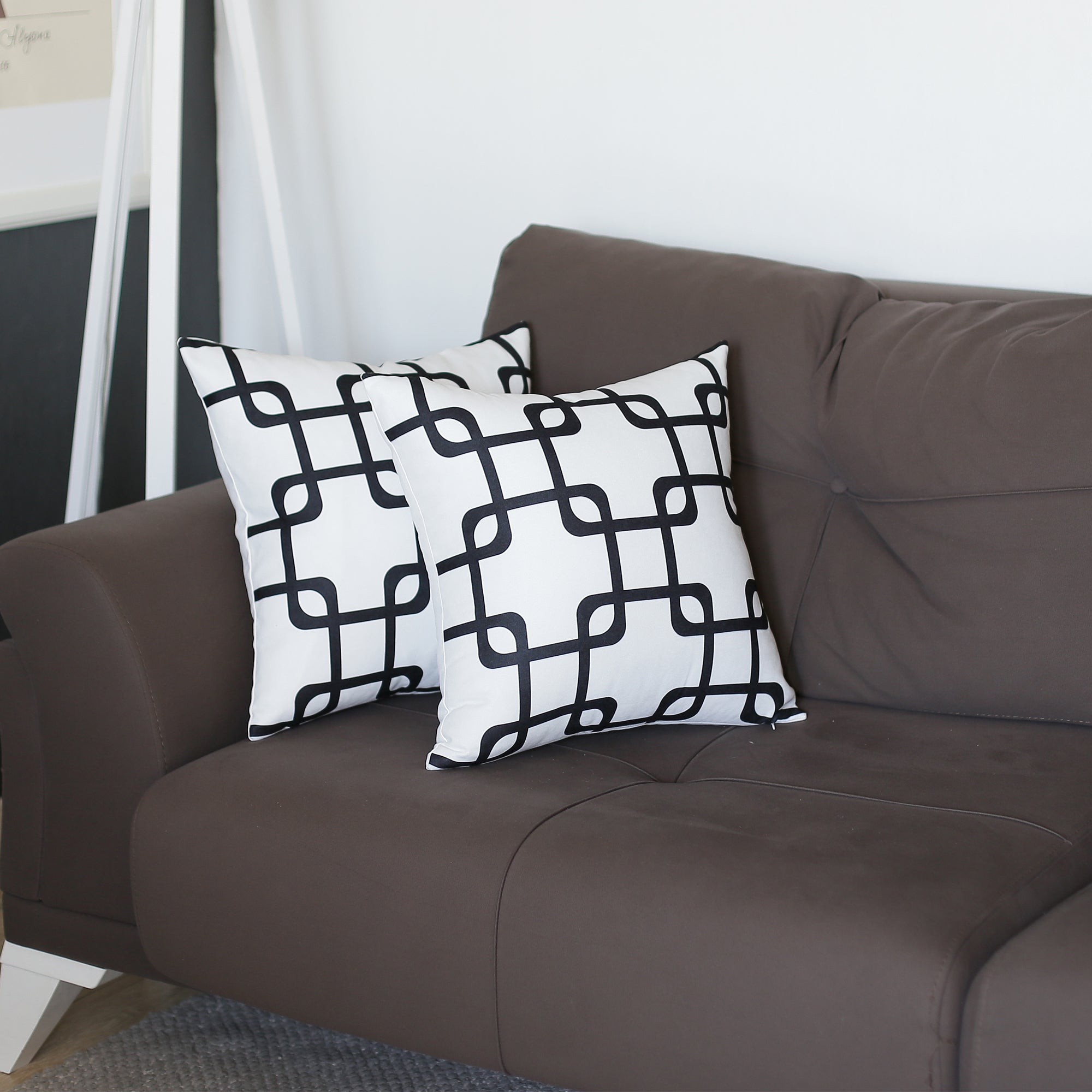 Two Geometric Black Squares 18" throw pillow covers rest on a dark brown couch in a living room setting, offering the convenience of hidden zipper closures for easy maintenance.