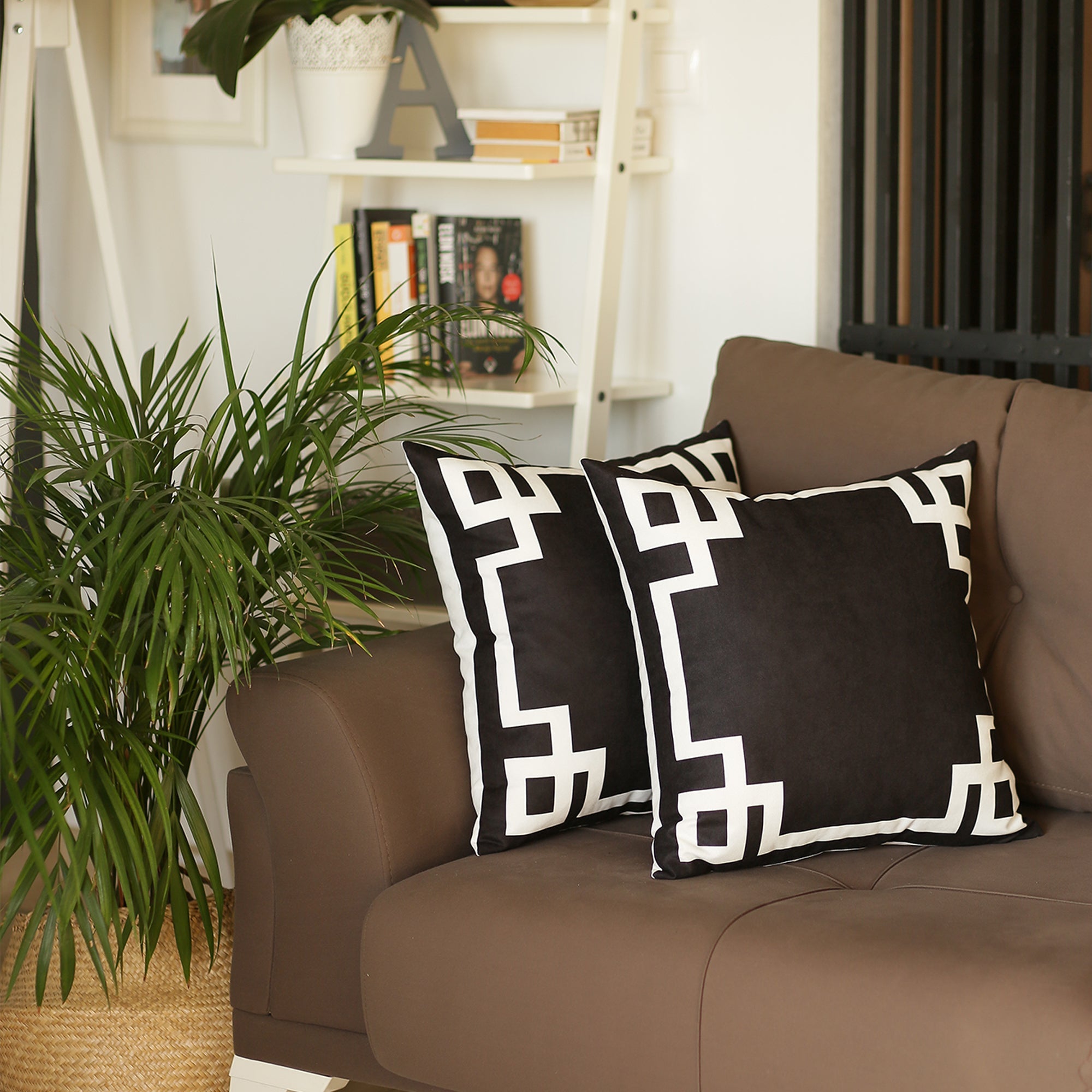 A brown sofa with Geometric Black&White Square Decorative Throw Pillow Covers (Set of 2) sits beside a plant. These pillow covers are designed with hidden zipper closures for easy maintenance. In the background, a white shelf displays a collection of books.