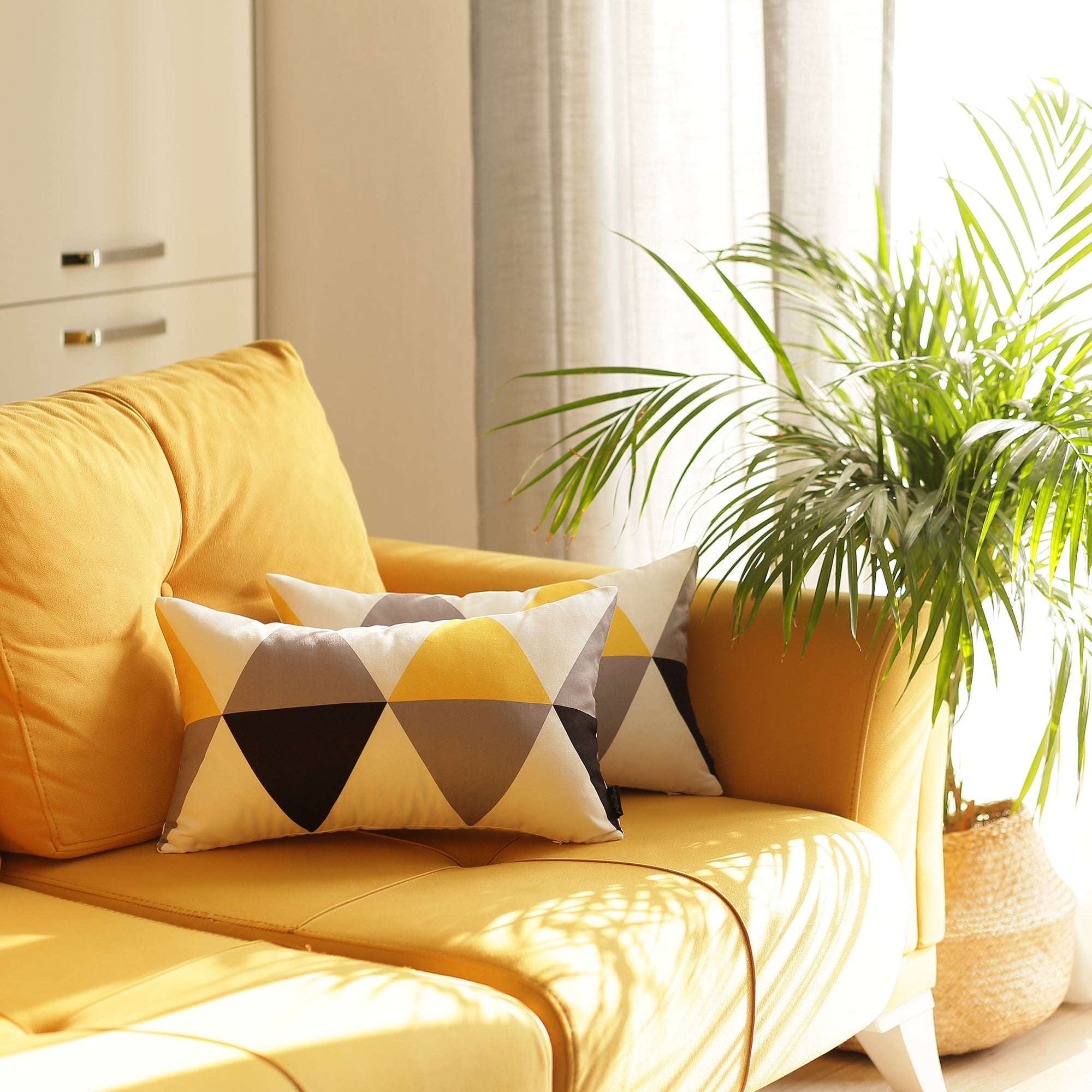 A yellow sofa, accessorized with two Scandi Yellow Gray Modern Lumbar Throw Pillow Covers, certified by OEKO-TEX Standard 100 and featuring a geometric pattern, is positioned beside a green potted plant and illuminated by natural light from a nearby window.