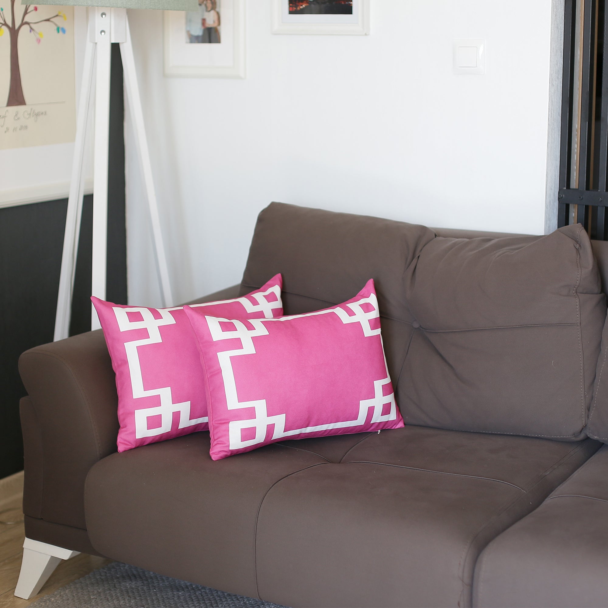 Adorning a brown sofa are two Geometric Pink&White Square 18" Throw Pillow Covers, each showcasing stylish geometric patterns. Paired with a green potted plant and a white bookshelf, these covers include convenient hidden zipper closures for effortless upkeep.