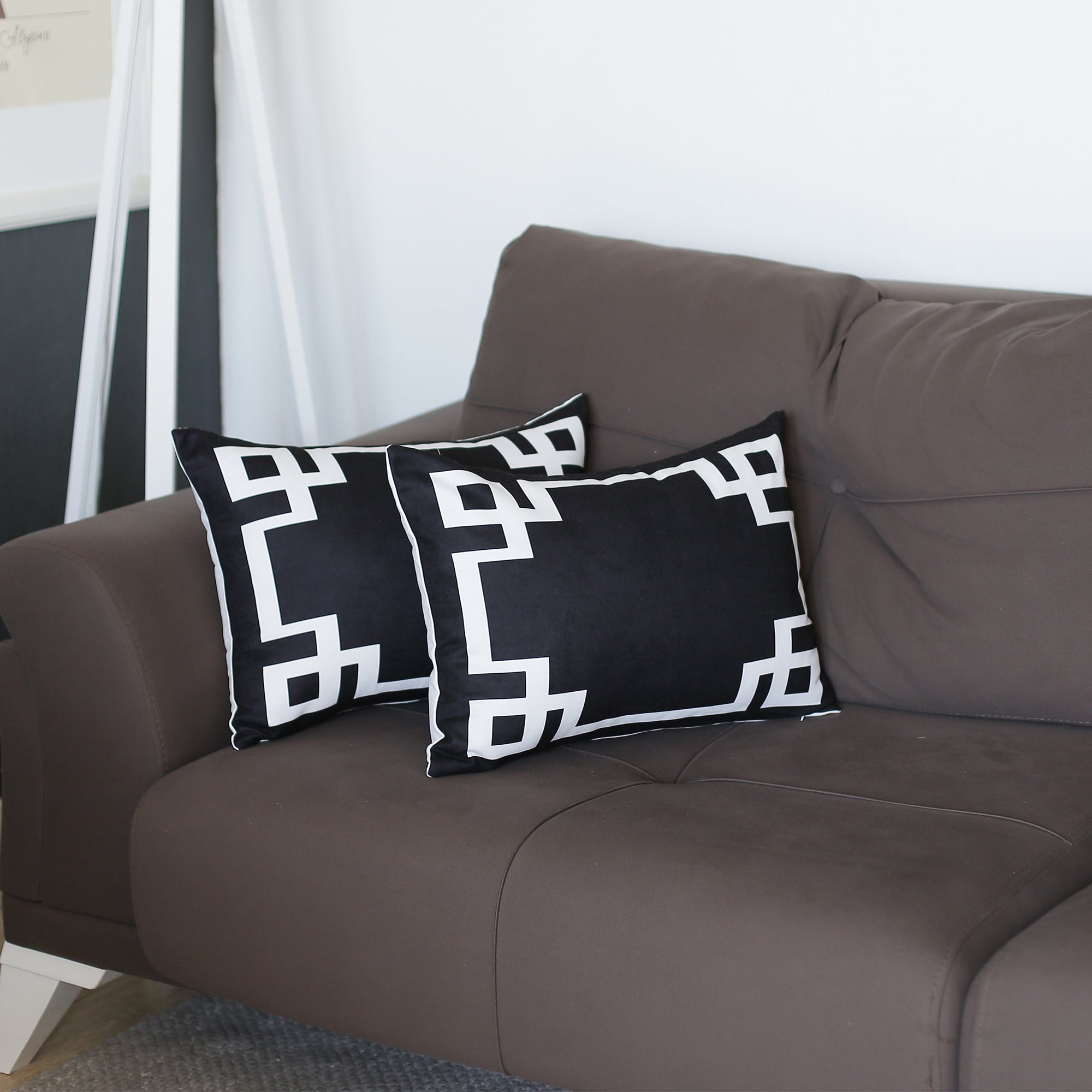 A brown sofa with Geometric Black&White Square Decorative Throw Pillow Covers (Set of 2) sits beside a plant. These pillow covers are designed with hidden zipper closures for easy maintenance. In the background, a white shelf displays a collection of books.