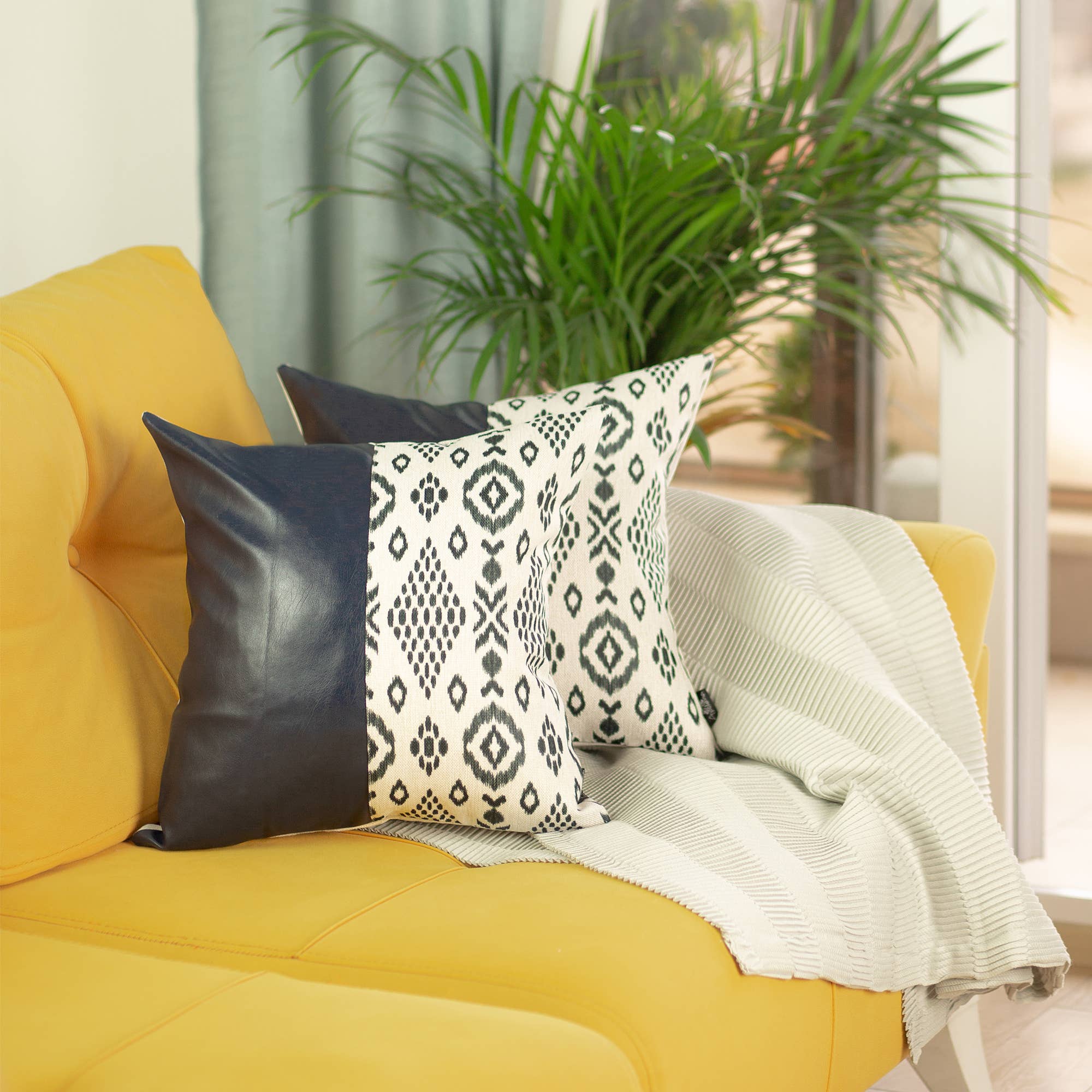 A Boho set of 2 handcrafted decorative throw pillow covers made of vegan faux leather with geometric squares complements a yellow sofa in a sunlit room, enhanced by a striped white blanket and a vibrant potted plant.