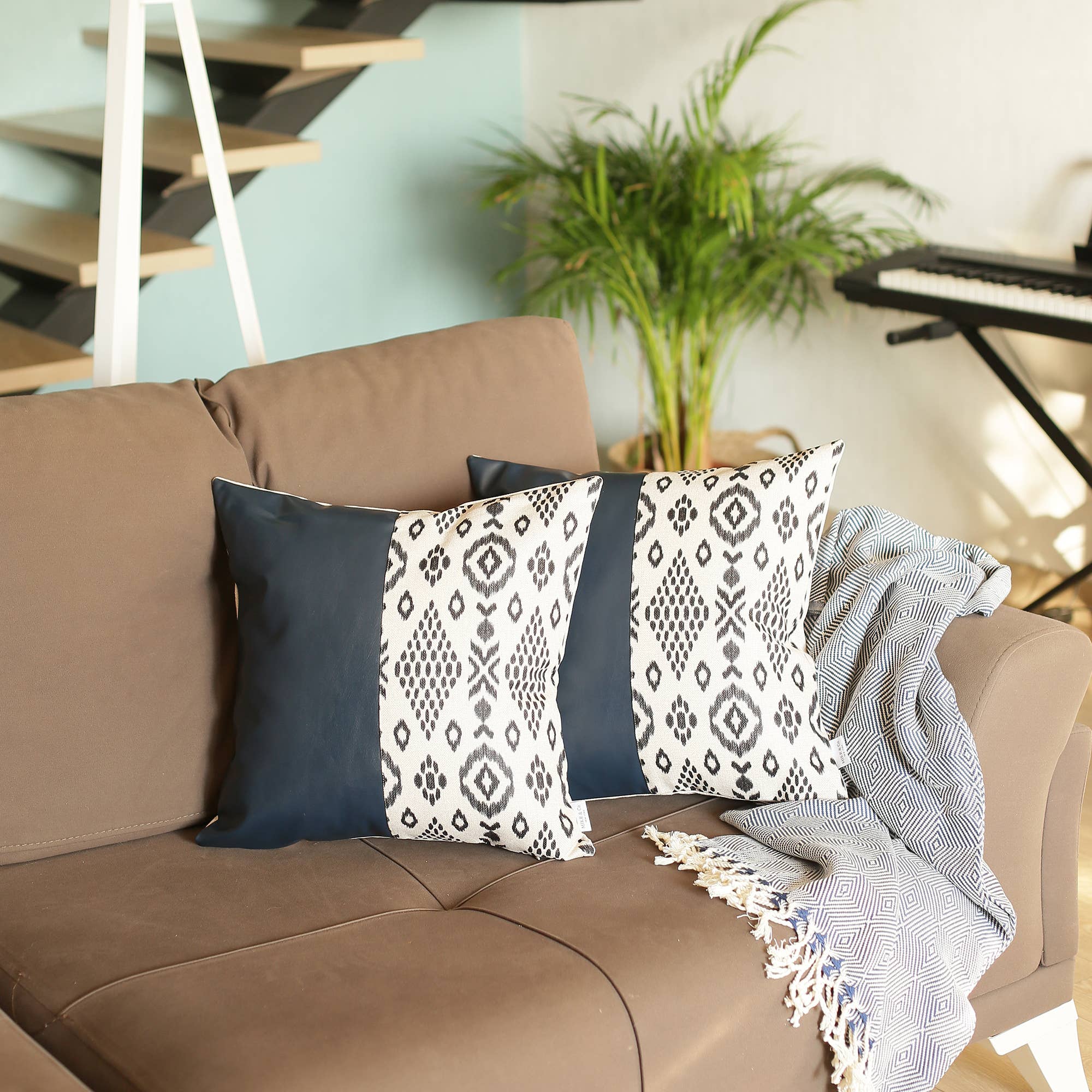 A Boho set of 2 handcrafted decorative throw pillow covers made of vegan faux leather with geometric squares complements a yellow sofa in a sunlit room, enhanced by a striped white blanket and a vibrant potted plant.