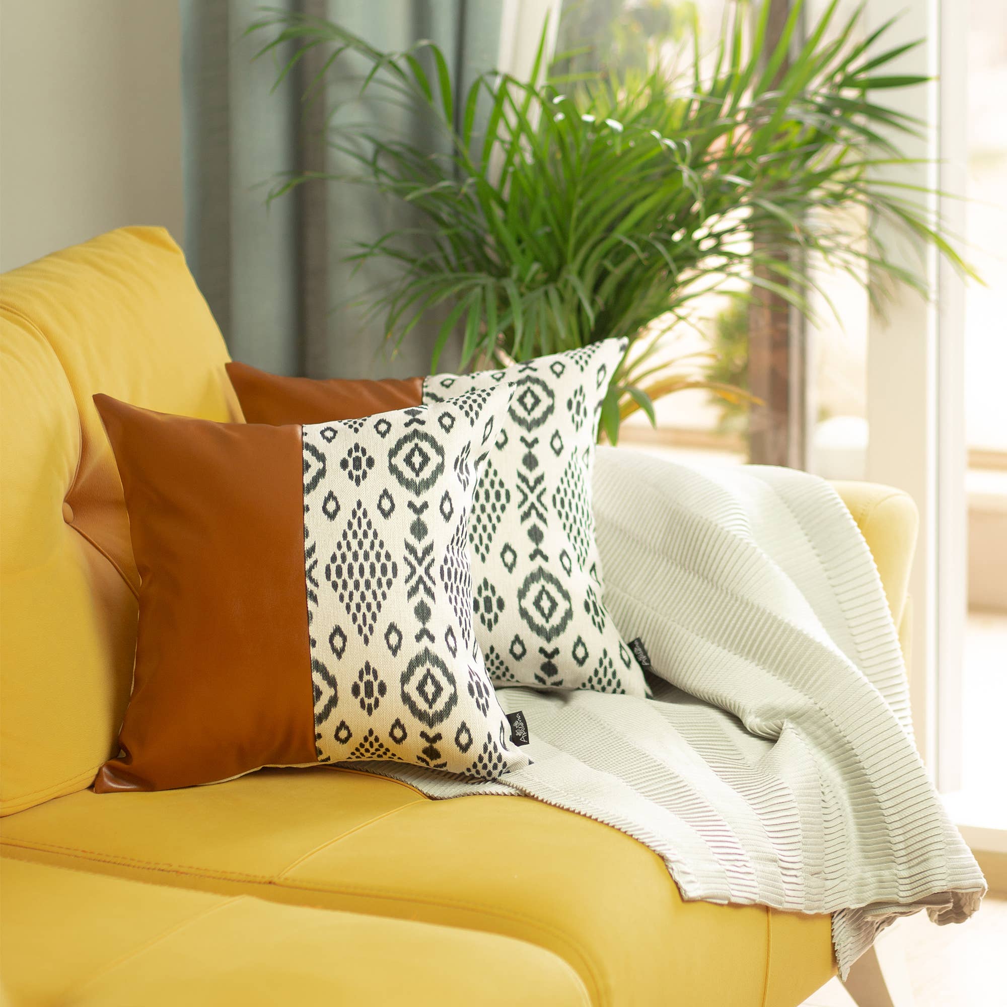 A Boho set of 2 handcrafted decorative throw pillow covers made of vegan faux leather with geometric squares complements a yellow sofa in a sunlit room, enhanced by a striped white blanket and a vibrant potted plant.