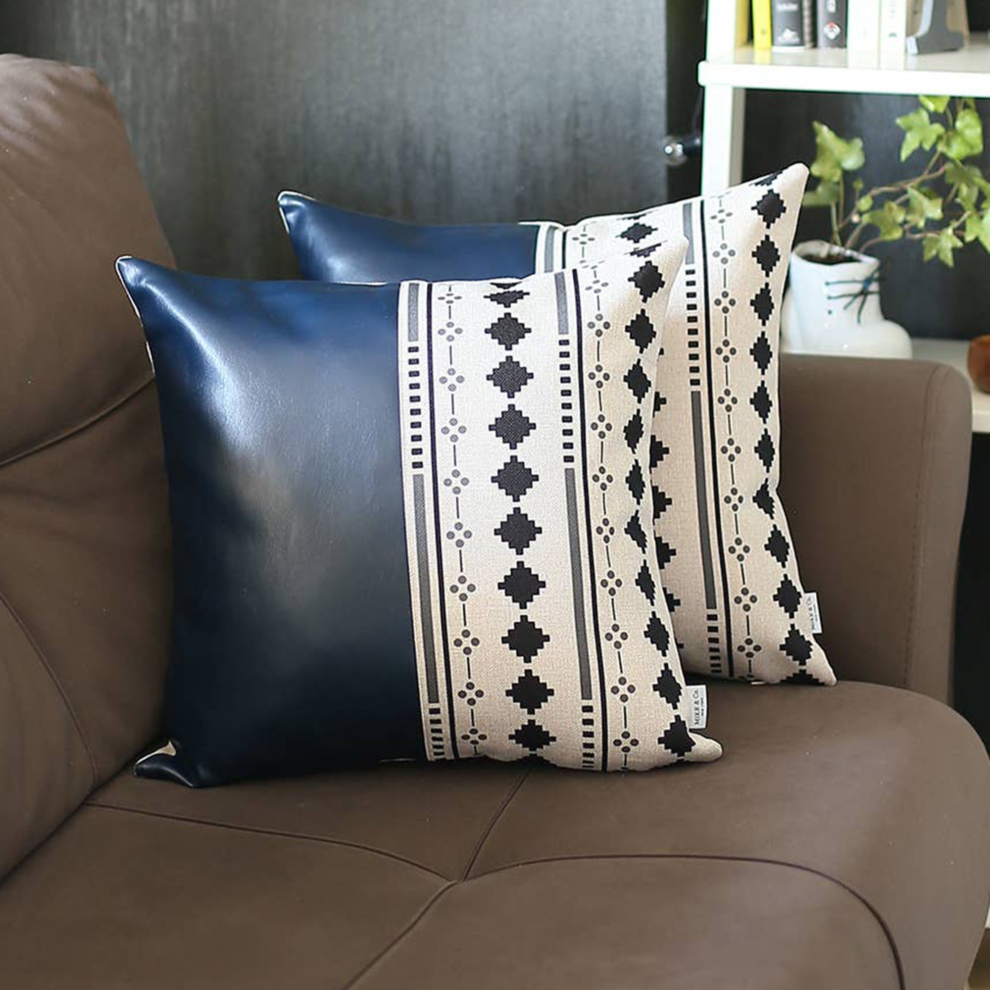 A pair of decorative pillows, featuring one in orange and another with a geometric design, are placed on a gray sofa in a modern living room. The Boho Set of 2 Handcrafted Decorative Throw Pillow Covers, made from vegan faux leather with geometric patterns, add an artistic flair that enhances the chic and cozy feel of the space.
