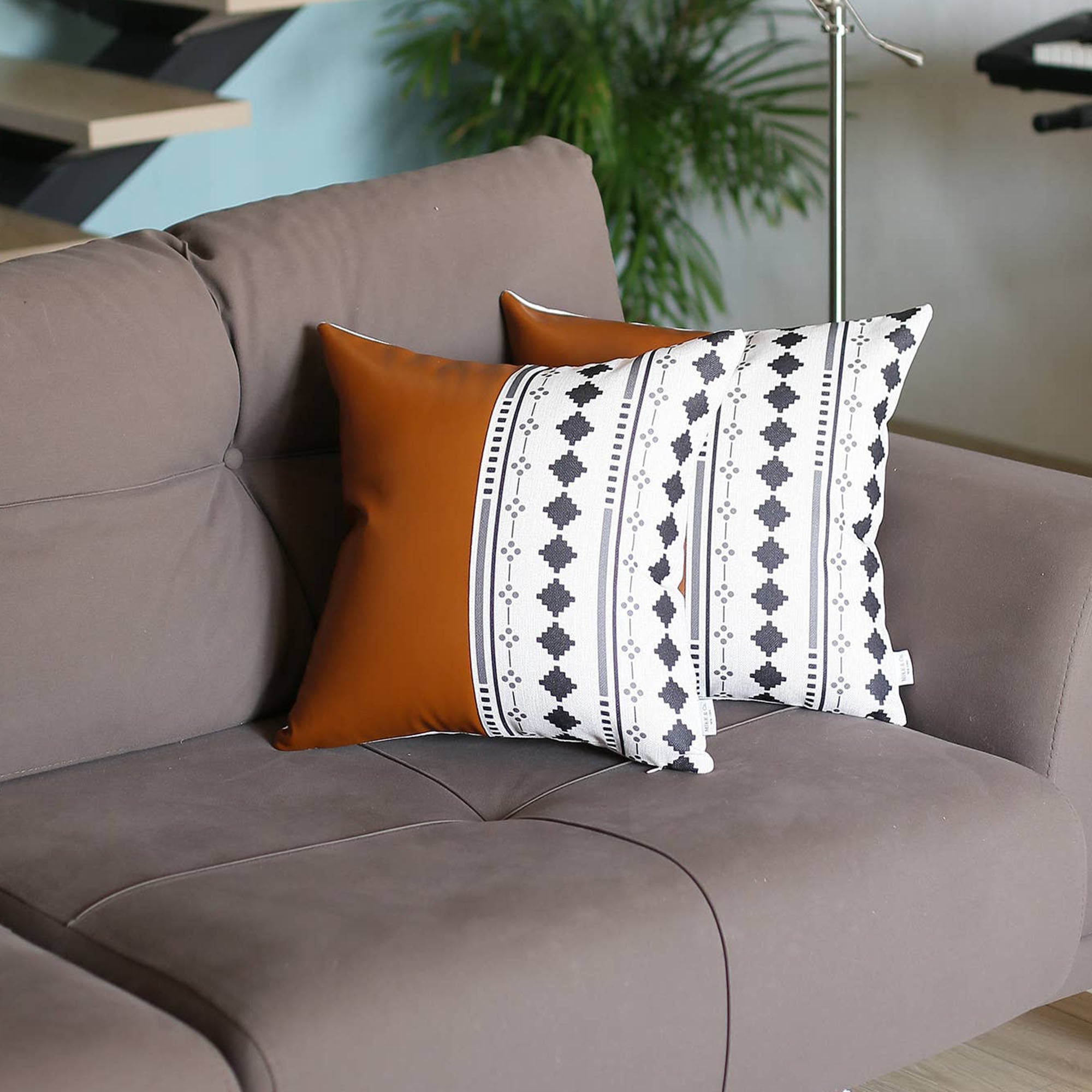 A pair of decorative pillows, featuring one in orange and another with a geometric design, are placed on a gray sofa in a modern living room. The Boho Set of 2 Handcrafted Decorative Throw Pillow Covers, made from vegan faux leather with geometric patterns, add an artistic flair that enhances the chic and cozy feel of the space.