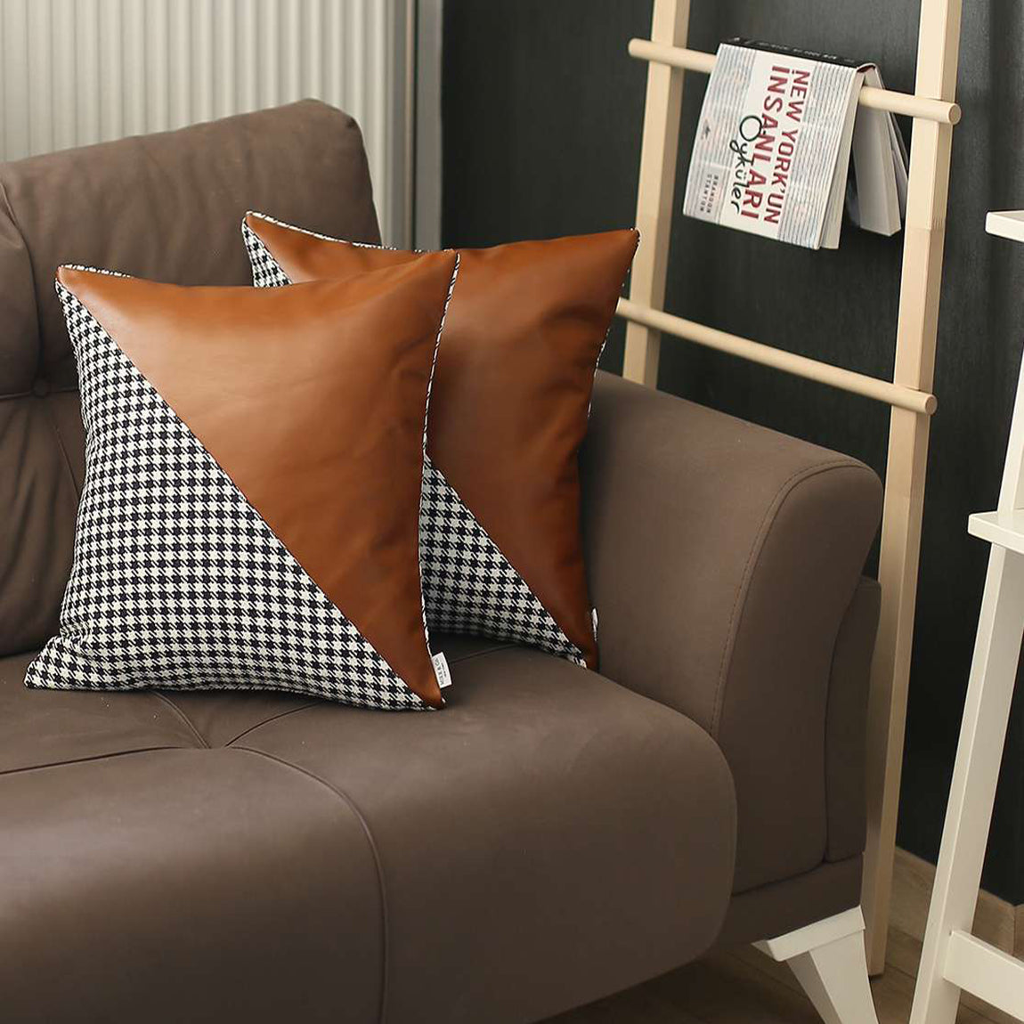 Two boho-chic 18" x 18" square throw pillow covers, handcrafted from vegan faux leather and featuring houndstooth patterns, adorn a brown couch. In the backdrop, a wooden ladder shelf holding a book enhances the cozy atmosphere with its harmonious blend of textures and eclectic design sensibilities.