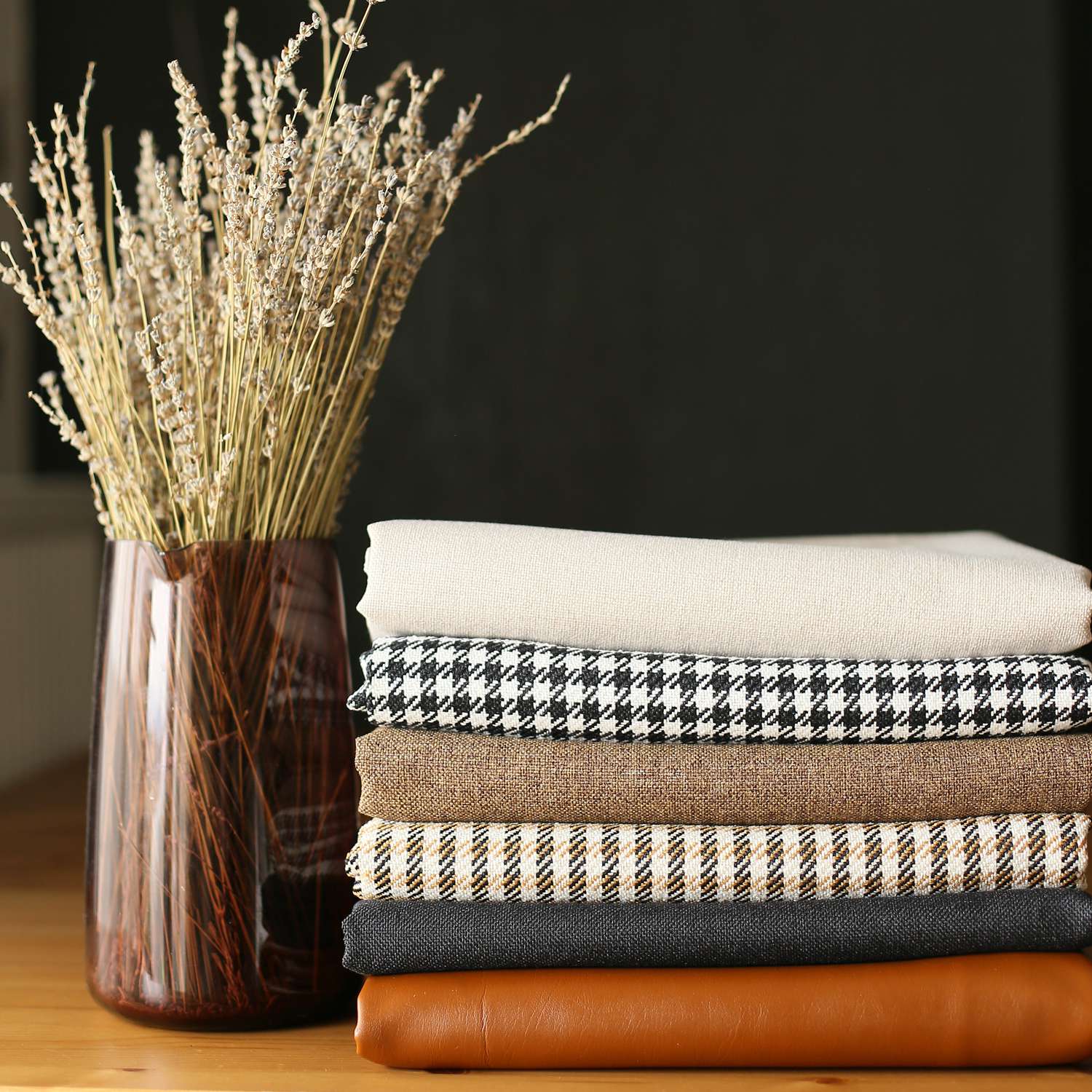 Two boho-chic 18" x 18" square throw pillow covers, handcrafted from vegan faux leather and featuring houndstooth patterns, adorn a brown couch. In the backdrop, a wooden ladder shelf holding a book enhances the cozy atmosphere with its harmonious blend of textures and eclectic design sensibilities.