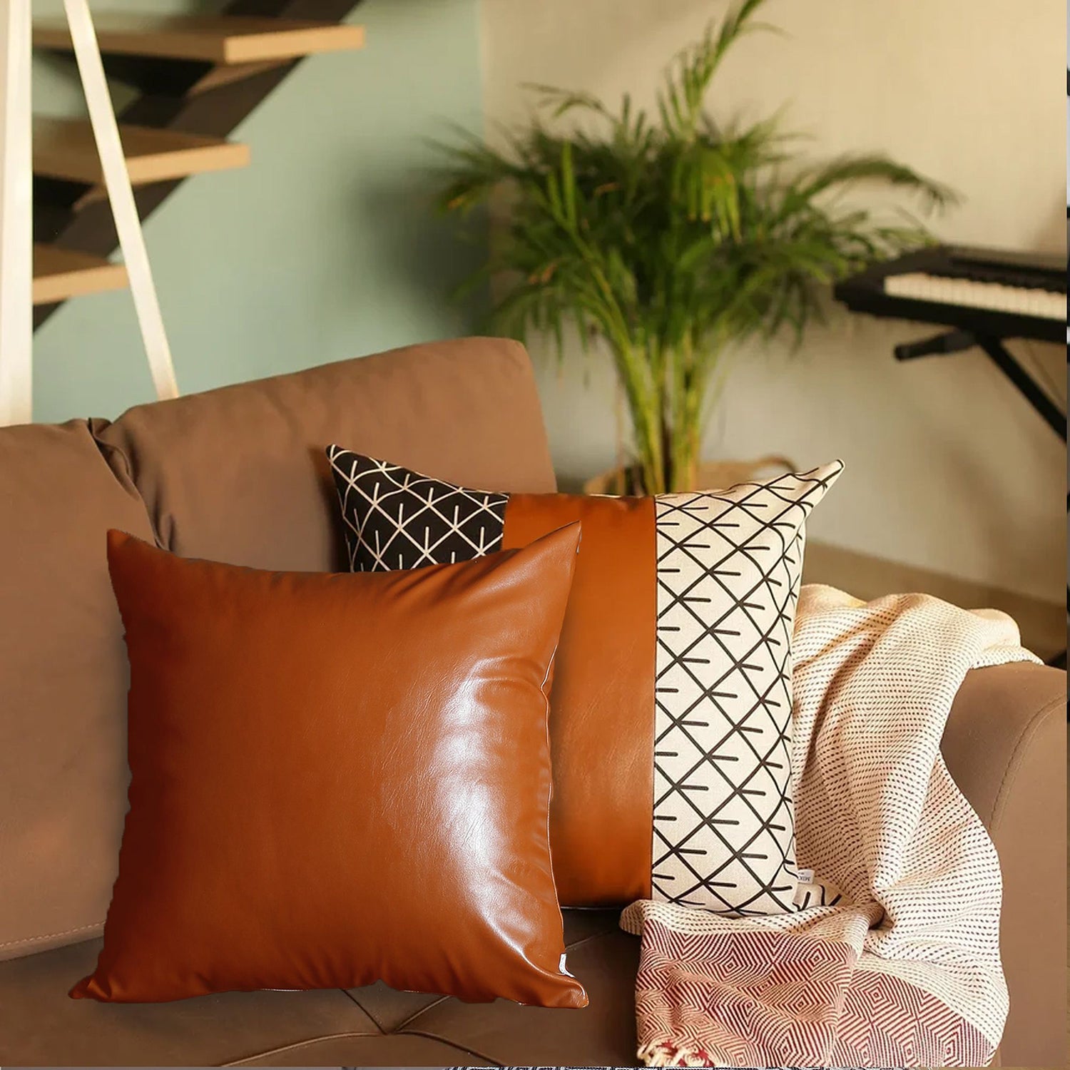 A brown couch decorated with a Bohemian Mixed Set of 2 Vegan Faux Leather Brown Geometric Throw Pillow Covers, enhanced by a knitted blanket and a plant in the background.