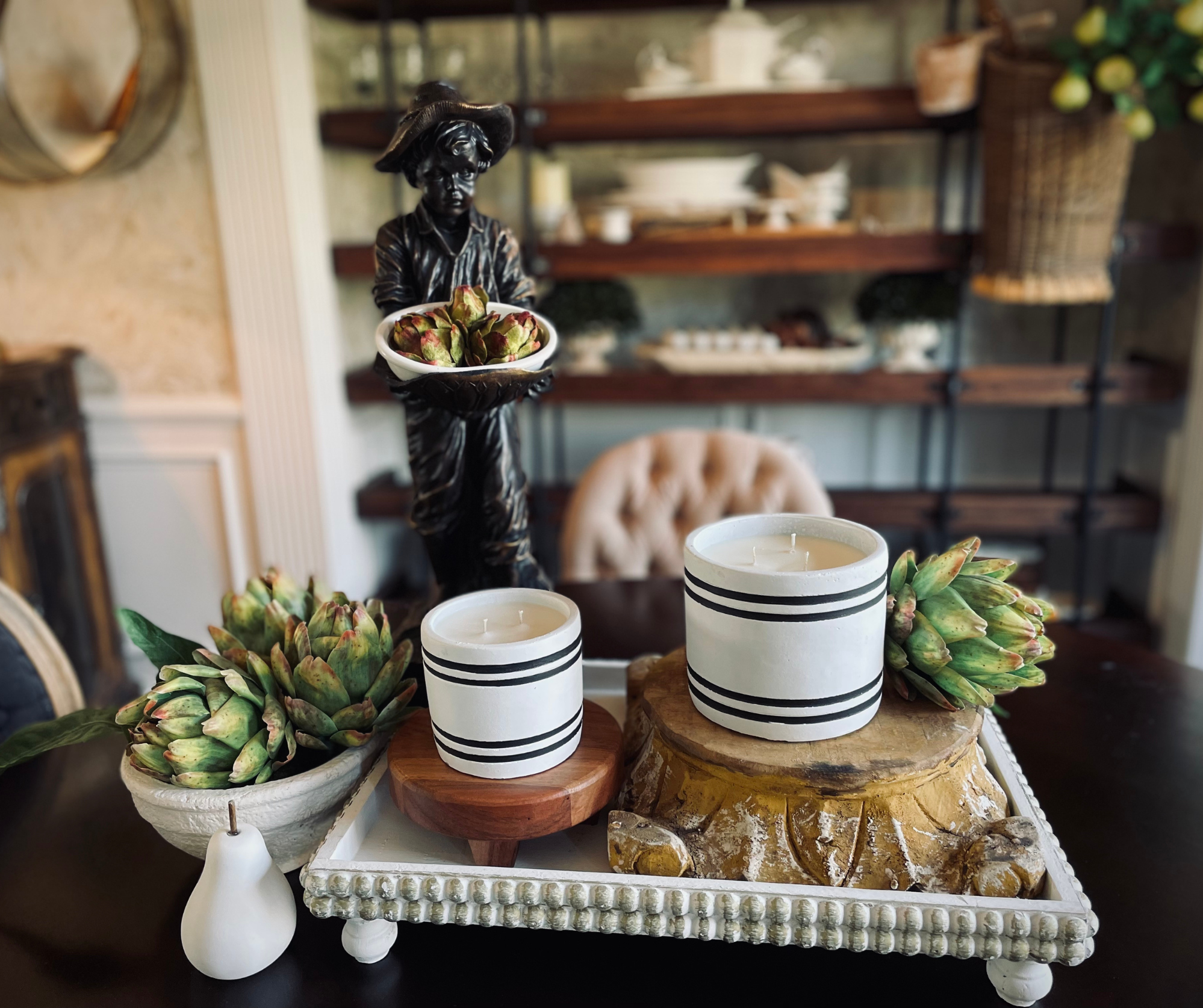 A hand-poured Luxury Small Striped Stone Designer Candle in Rosemary & Mint fragrance, featuring black stripes and placed on a wooden stand, is enhanced by surrounding greenery and vibrant red-orange fruits. Crafted from natural soy wax.