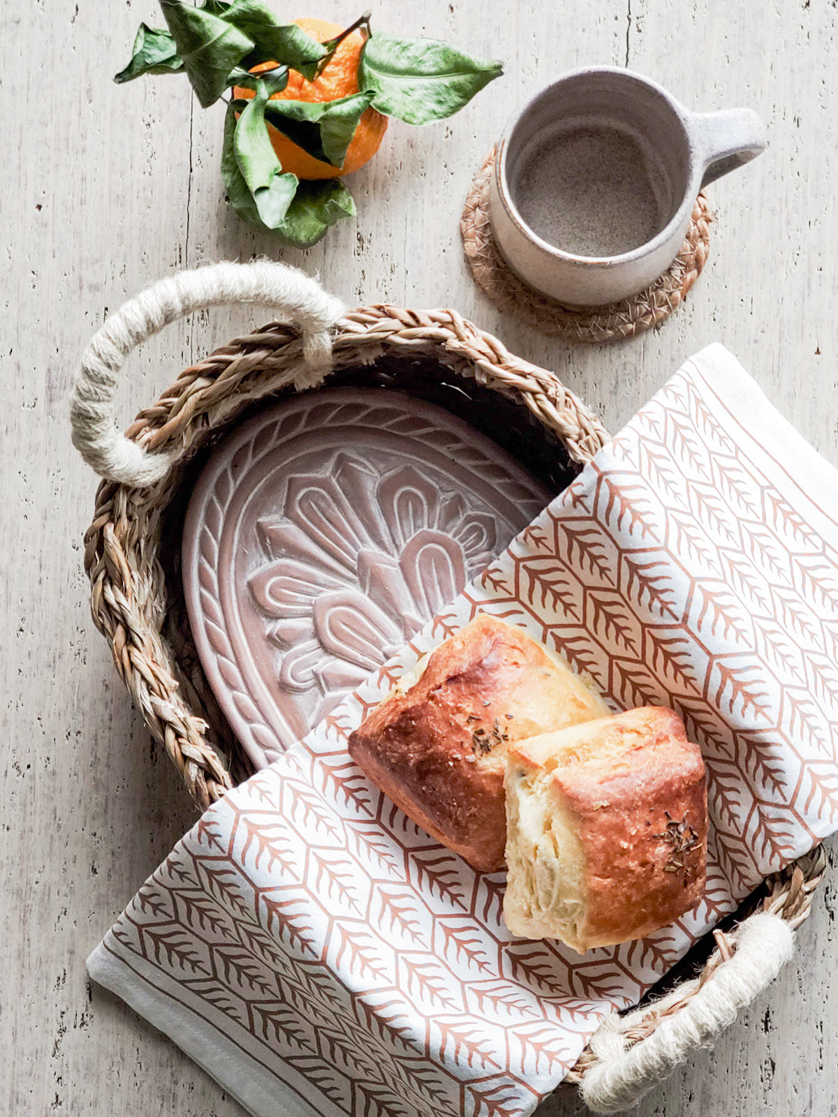 A decorative ceramic plate is beautifully set in a handwoven seagrass basket featuring handles, called the Bread Warmer & Basket Gift Set with Tea Towel - Flower. Below the basket are two patterned towels, crafted by Fair Trade artisans, showcasing leaf designs in black and orange.