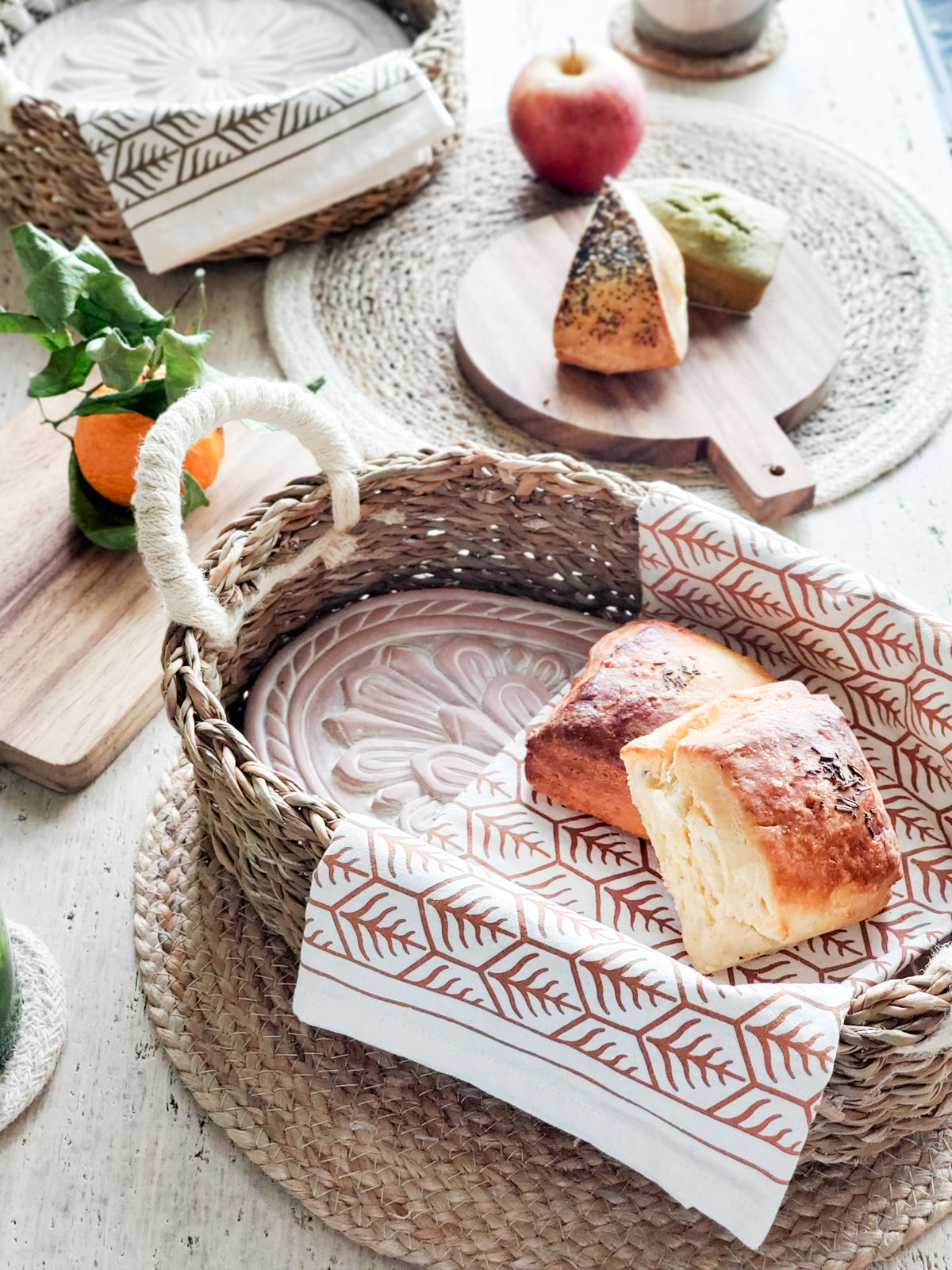 A decorative ceramic plate is beautifully set in a handwoven seagrass basket featuring handles, called the Bread Warmer & Basket Gift Set with Tea Towel - Flower. Below the basket are two patterned towels, crafted by Fair Trade artisans, showcasing leaf designs in black and orange.