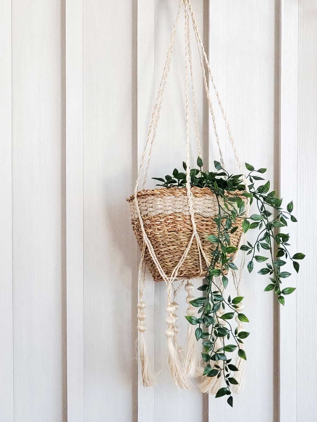 The Plant Hanger - Fiora is a beautiful handmade macramé plant holder, showcasing a black pot adorned with a beige jute rope design, complemented by wooden beads and tassels. Crafted by Fair Trade artisans, it represents sustainable handicraft.