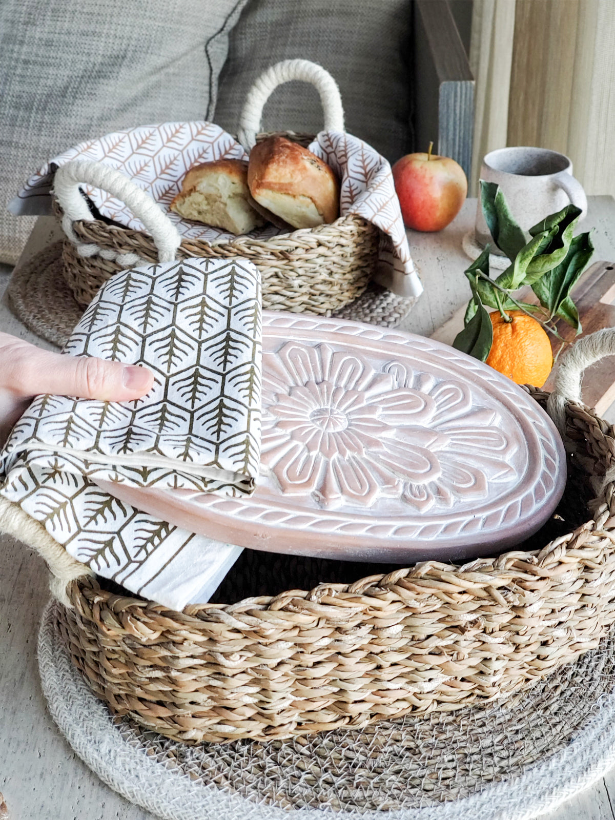 A decorative ceramic plate is beautifully set in a handwoven seagrass basket featuring handles, called the Bread Warmer & Basket Gift Set with Tea Towel - Flower. Below the basket are two patterned towels, crafted by Fair Trade artisans, showcasing leaf designs in black and orange.