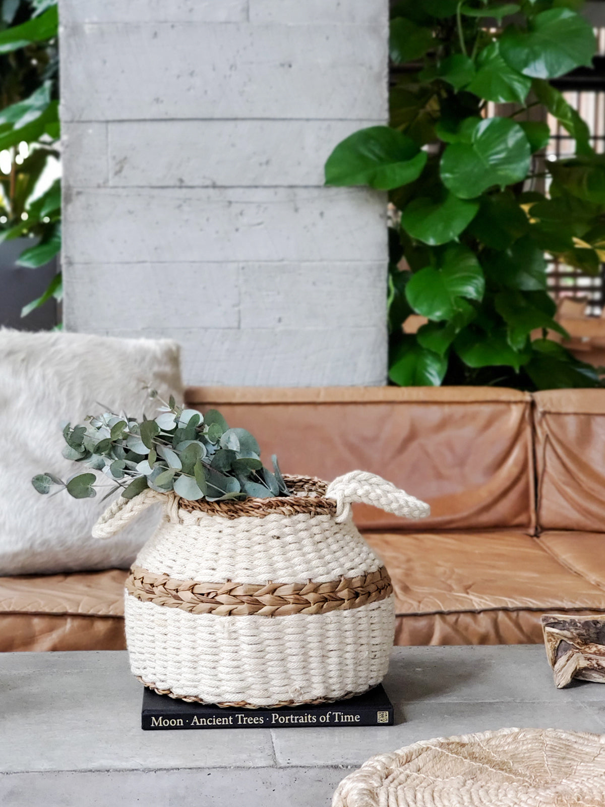 The Ula Blanco Basket is a handwoven piece with two handles, showcasing a natural color and a brown accent stripe around the middle, crafted by sustainable artisans using ethically sourced materials.